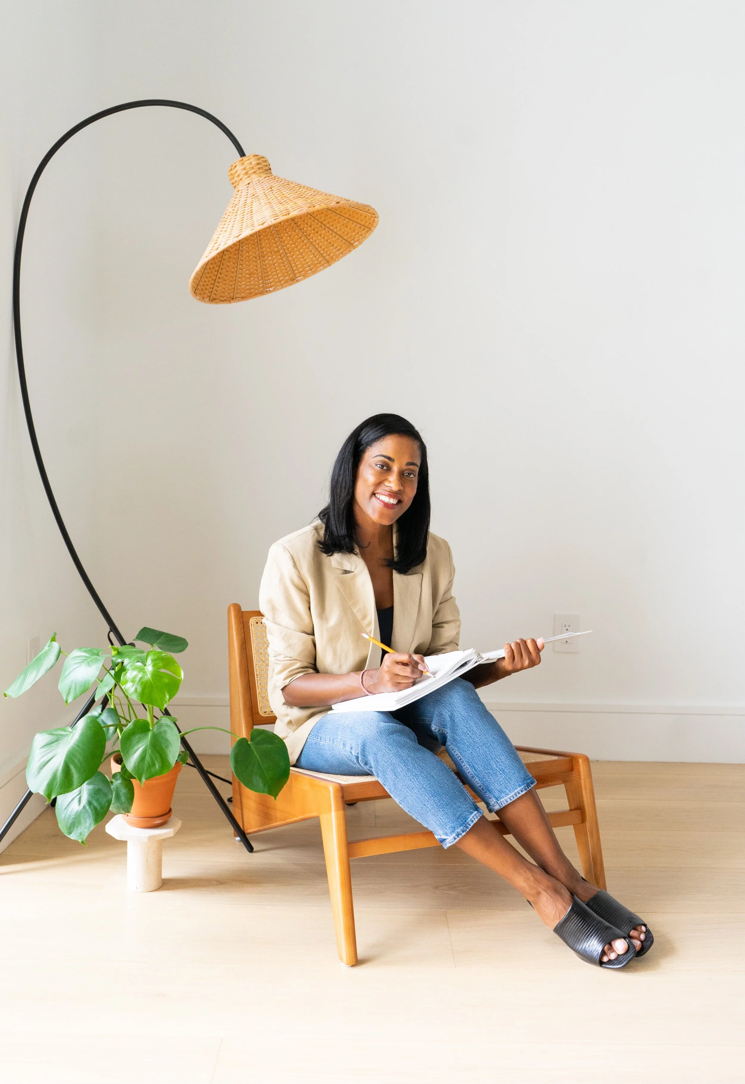 A woman with black hair sitting on a wooden chair, smiling, holding a notebook and pencil, wearing a beige blazer, blue jeans, and black open-toe slippers, near a green potted plant, with a large white wall in the background and a tall arc floor lamp overhead.