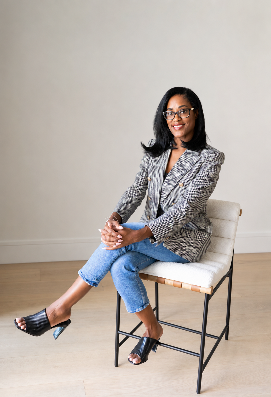 A woman sitting on a white chair with a light beige wall in the background, wearing a checkered blazer, blue jeans, and high-heeled shoes, smiling at the camera.