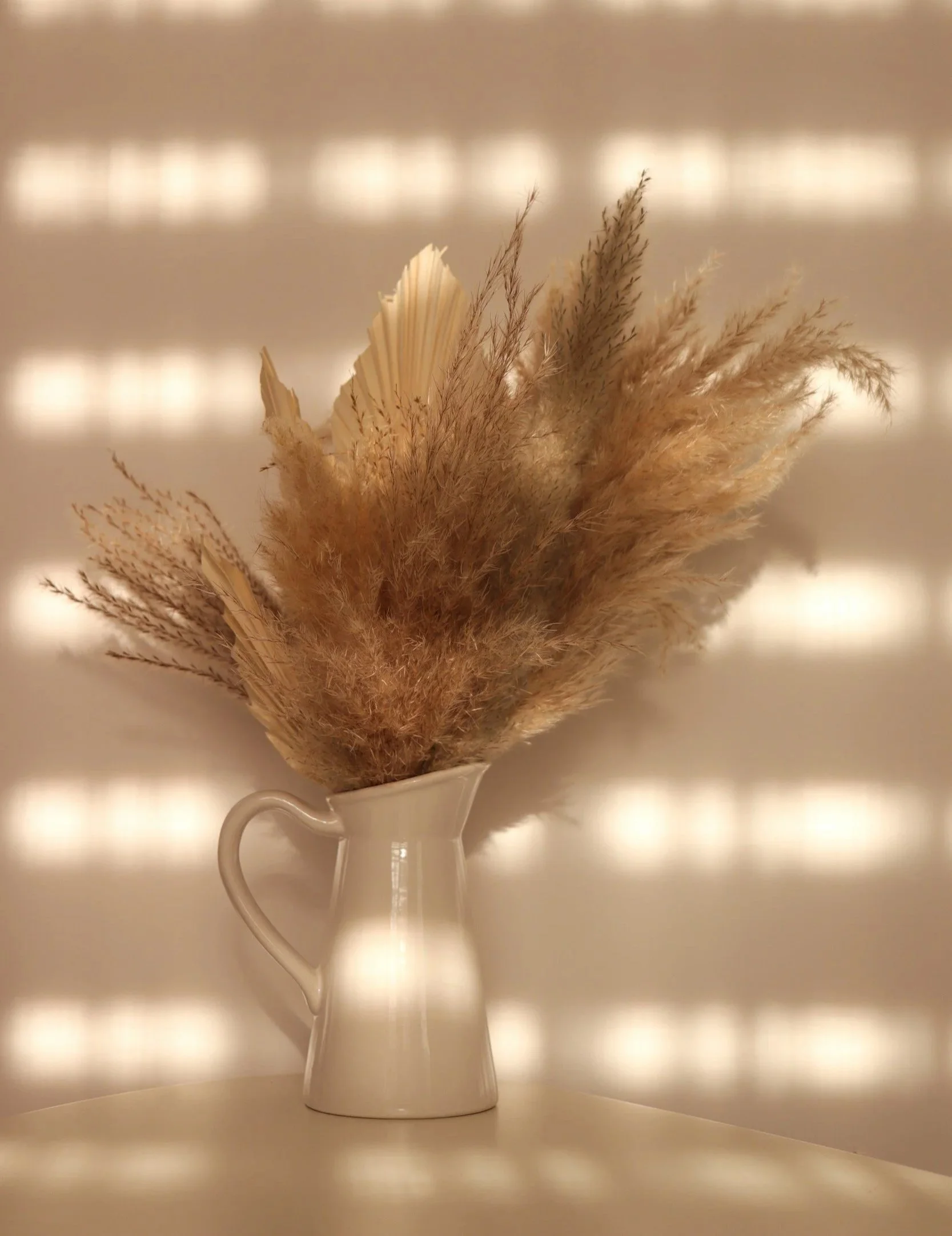 A white ceramic pitcher holding dried pampas grass and other dried foliage, placed against a neutral background with soft, reflective lighting.