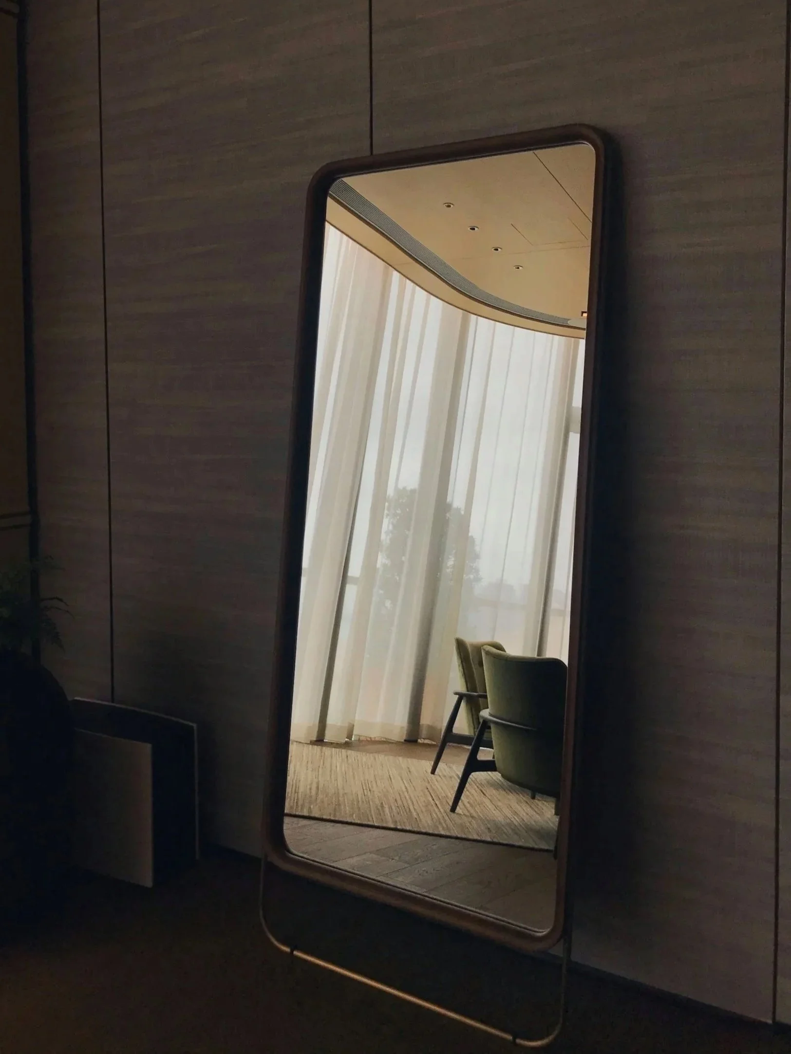 A large full-length mirror with rounded edges leaning against a wall, reflecting a room with a curtain, a chair, and a beige carpet.