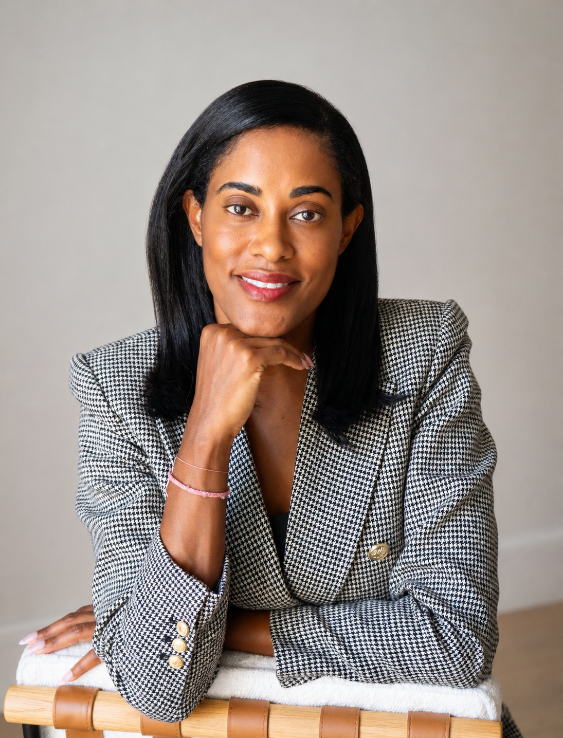 Portrait of a woman with shoulder-length black hair, wearing a houndstooth blazer, sitting with her chin resting on her hand, smiling against a plain background.