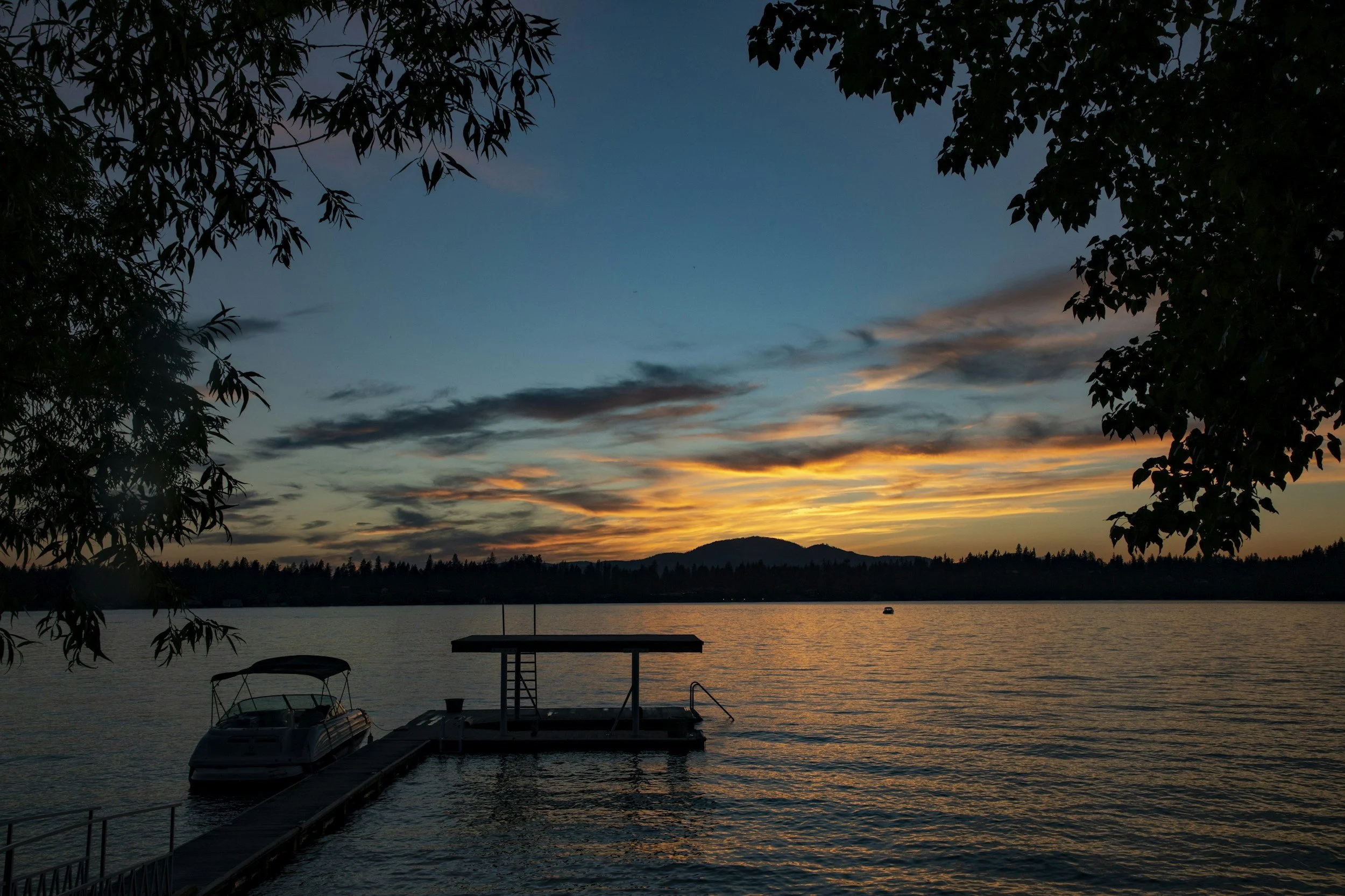 Sunset over a lake with boats docked at a pier, silhouetted trees on the sides, and mountains in the distance.