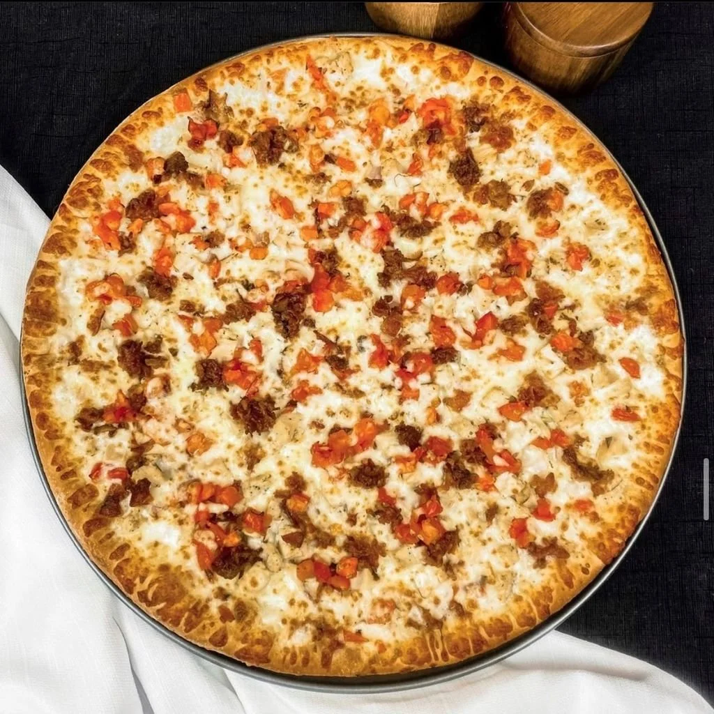 A large pizza with cheese, bacon, chicken, and chopped tomatoes on a round metal tray.