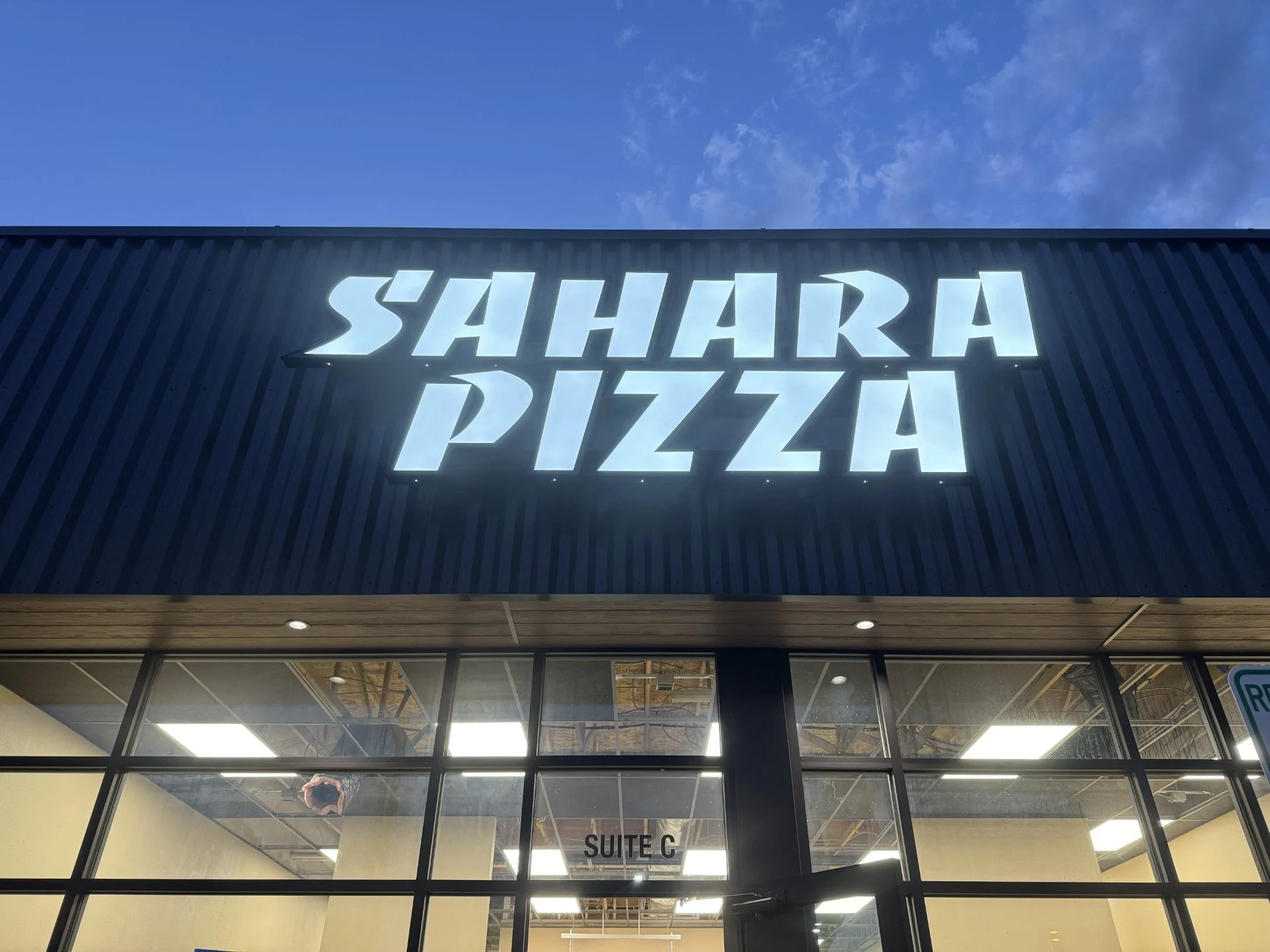 Exterior view of Sahara Pizza restaurant with large illuminated sign against a blue sky, glass windows showing the interior, and a sign indicating Suite C inside.