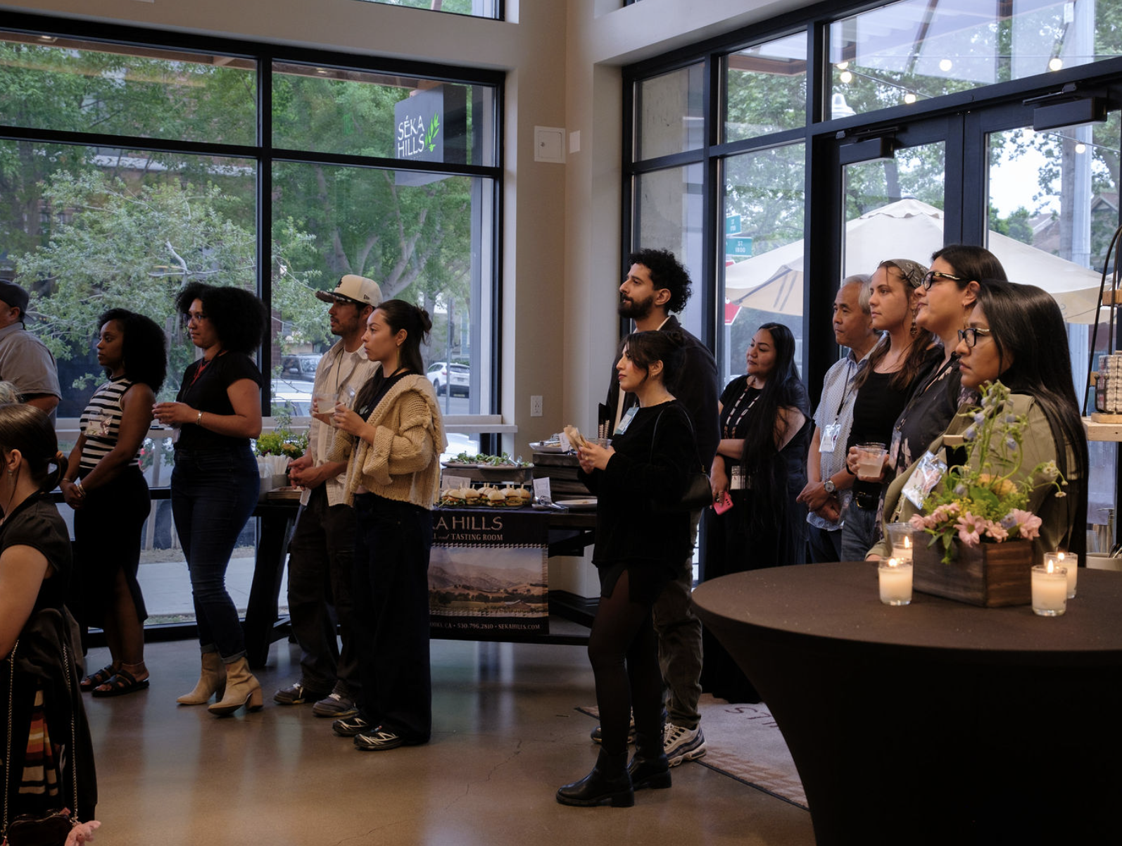Group of diverse people standing in a row at an indoor event, with a large window showing trees outside and a table with flowers and candles in the foreground.