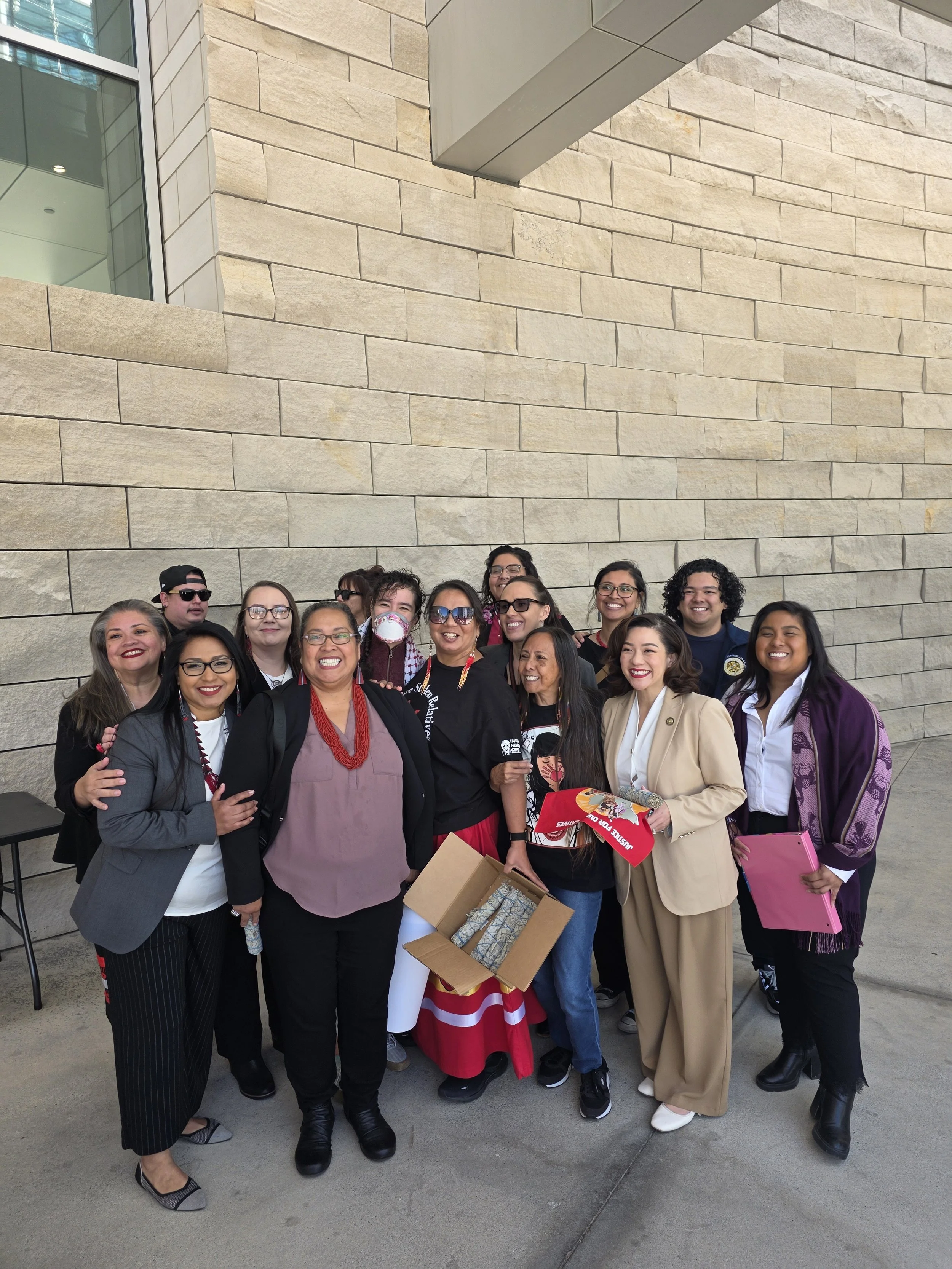 A diverse group of people standing together outside a building made of light-colored stone against a modern glass structure. They are smiling and appear to be celebrating or participating in a group event.