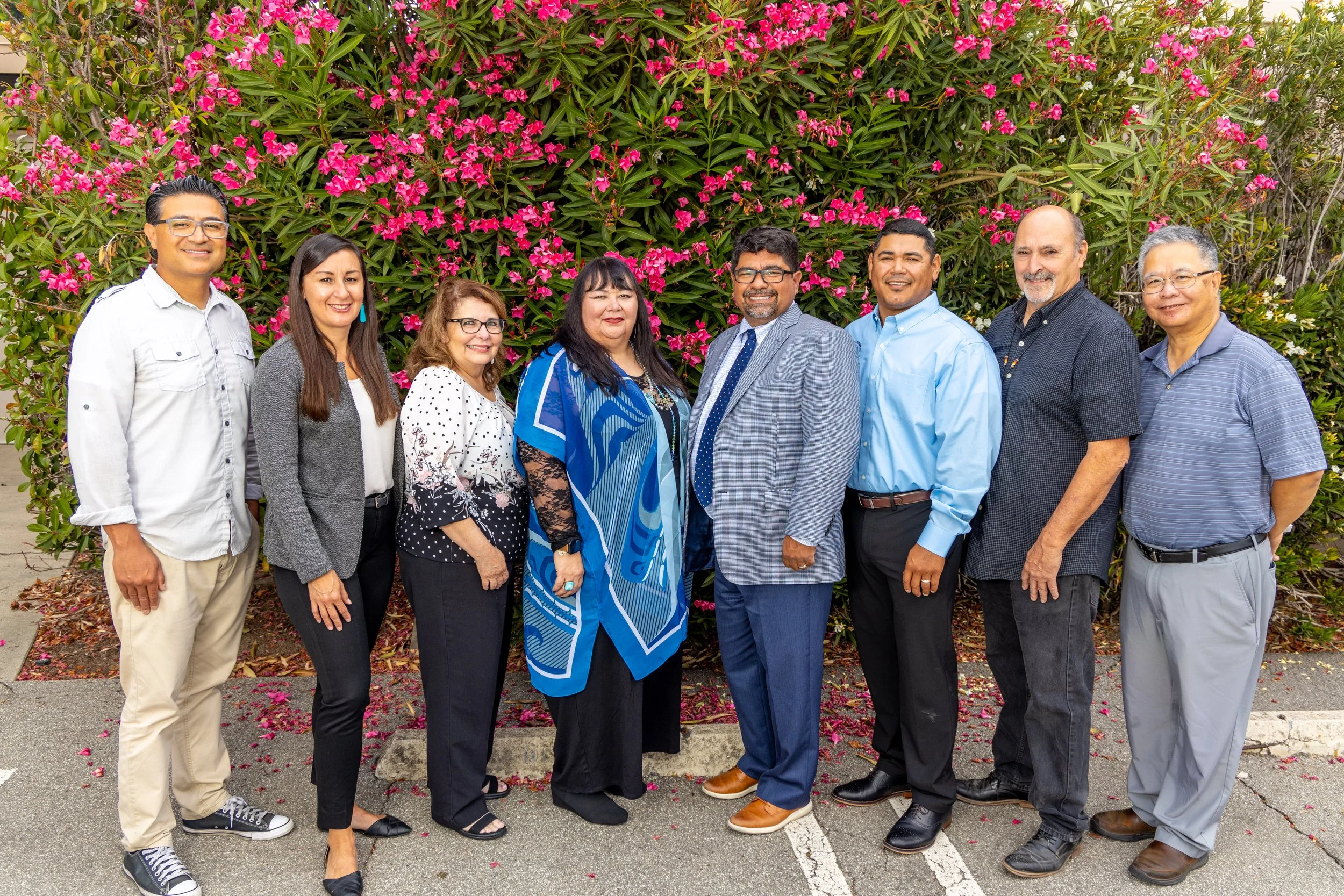 Group of nine diverse adults standing outdoors in front of a large pink flowering bush, dressed in business casual attire, smiling at the camera.