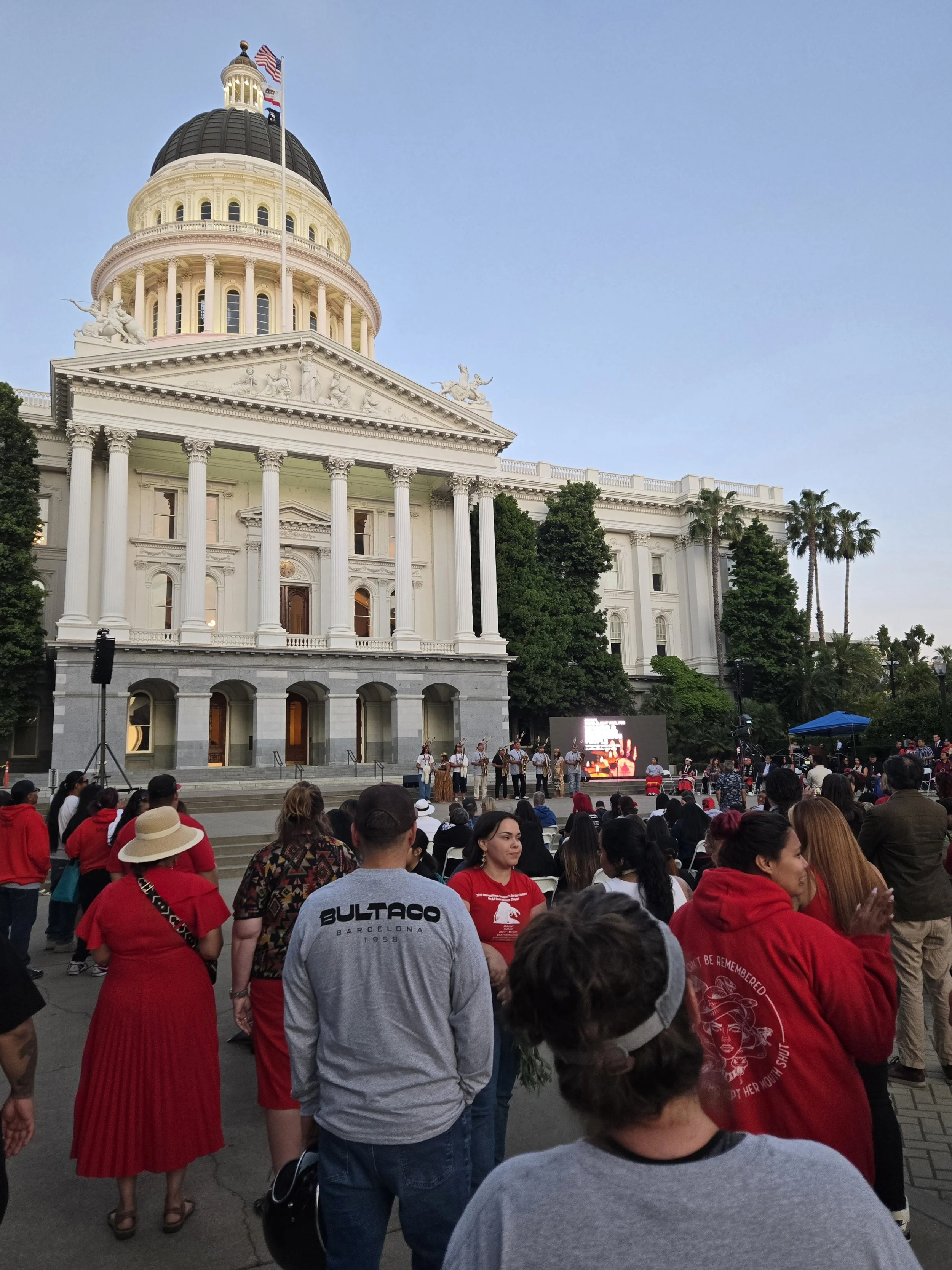 People gathered in front of the California State Capitol building, where a group is performing on a stage with a digital screen, while an audience is watching. The building has a prominent dome with flags on top and classical architecture.