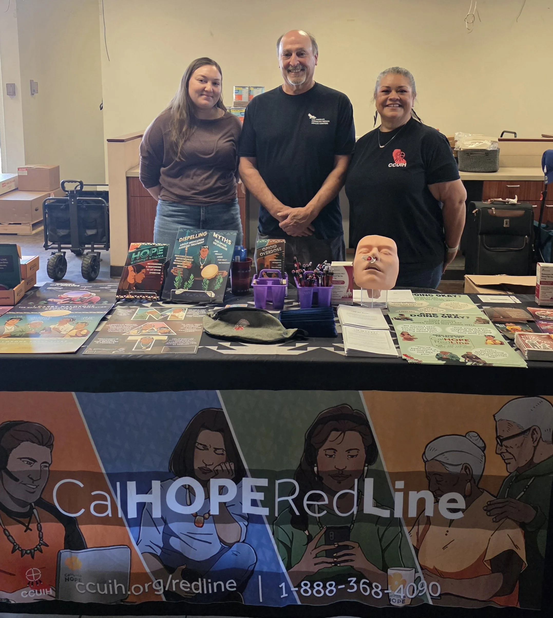 People standing behind a table with informational materials, including brochures and a CPR mannequin, at an outreach event. The table displays the URL ccu.org/redline and a phone number 1-888-368-4090. The event appears to focus on health or community outreach.