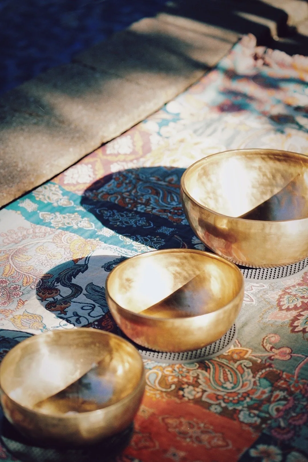 Three polished brass singing bowls resting on a colorful, patterned textile with a stone bench in the background.