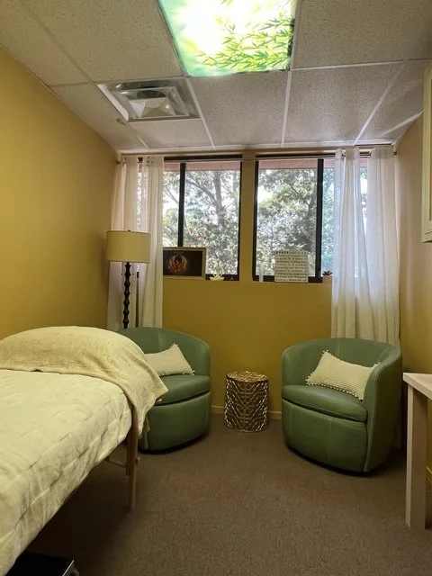 Small room with yellow walls, a bed, two green armchairs with pillows, a wicker side table, a window with white curtains, a lamp, and decorative items on the window sill.