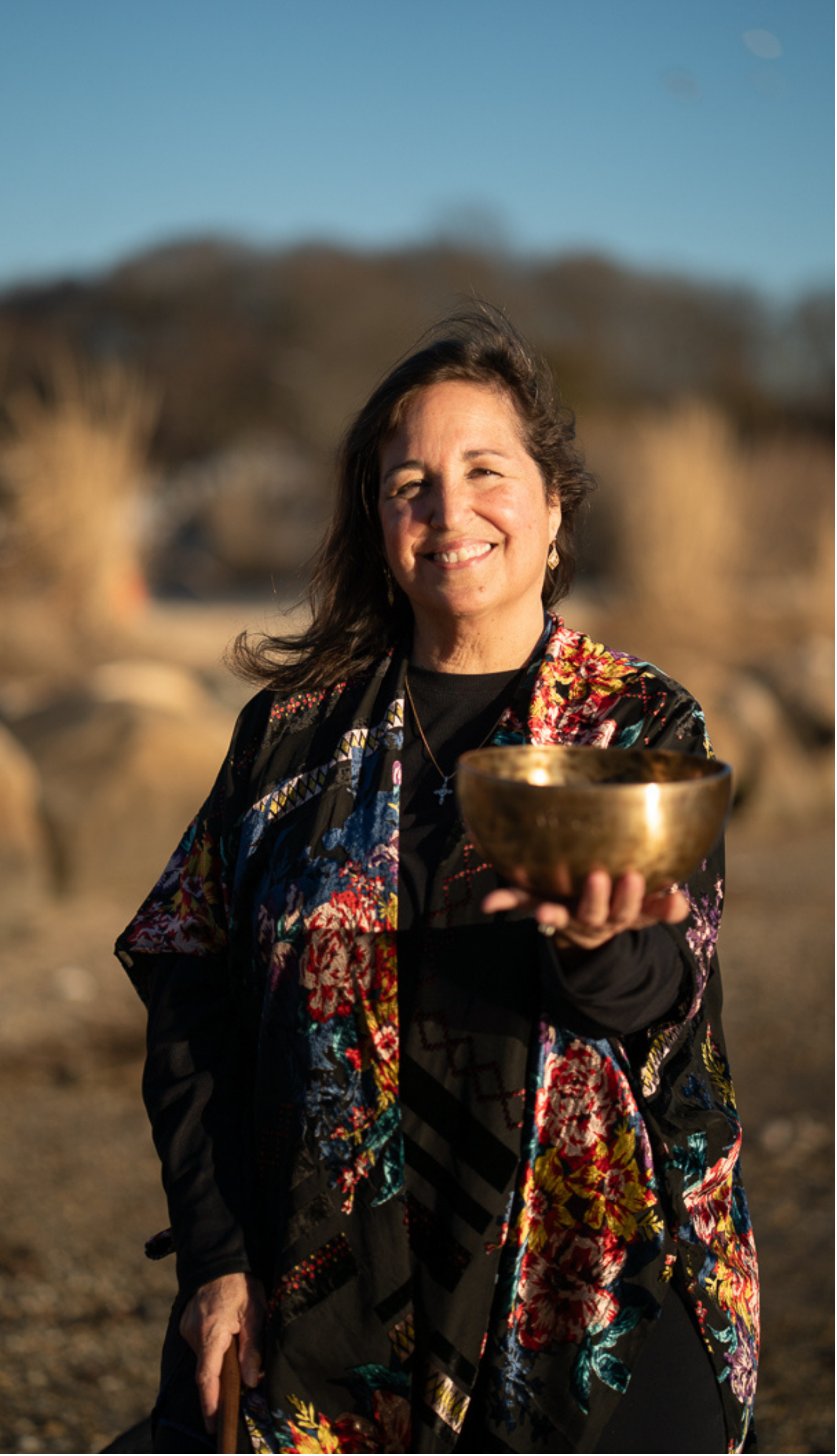 Woman smiling outdoors, holding a golden bowl, wearing a colorful embroidered jacket and earrings, with a blurred natural background.