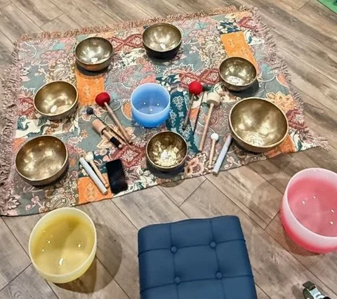 A padded mat sits diagonally just outside the picture surrounded by 7 bronze sound bowls, 2 crystal bowls with gong utensils sitting fanned out pointing towards the bowls. All bowls & utensils are sitting on a small quilted scarf on a wood floor.