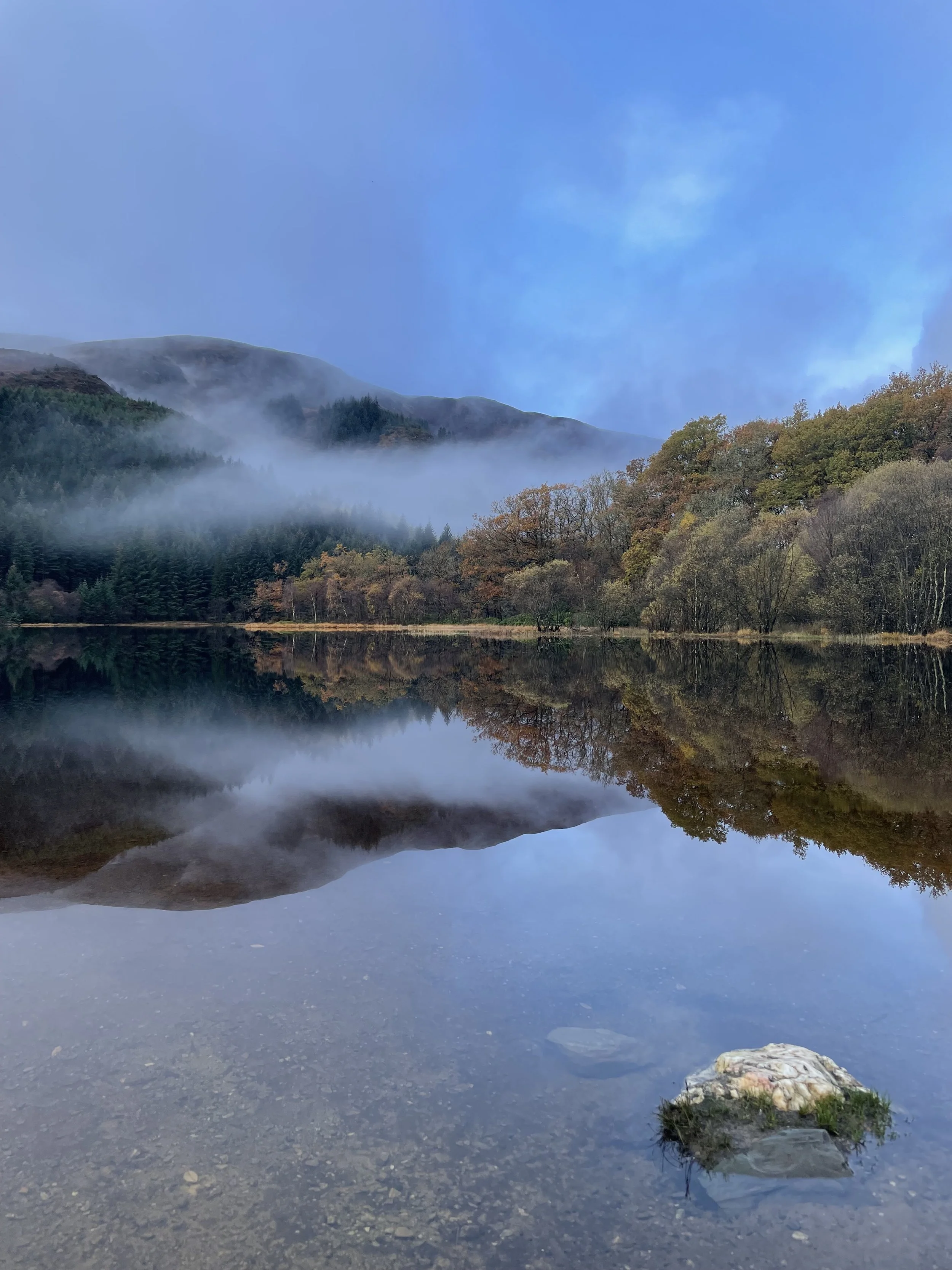 A calm lake reflecting a mountain range with mist, trees, and a blue sky with clouds.