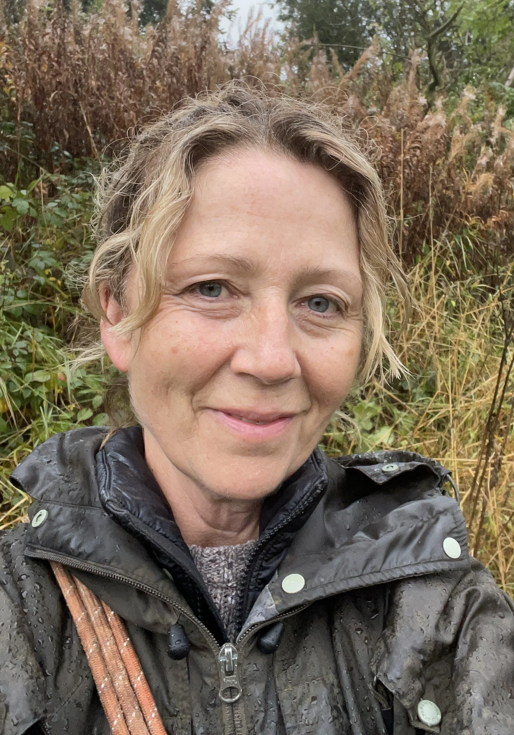 A woman with light skin, blue eyes, and curly blonde hair smiling outdoors in a forest with fall-colored foliage. She is wearing a black rain jacket with raindrops on it.