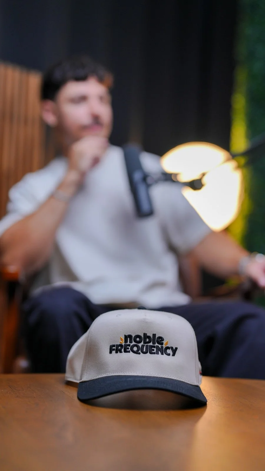 A Noble Frequency branded hat on the studio table, with a podcast guest seated behind it in Temecula–Murrieta.