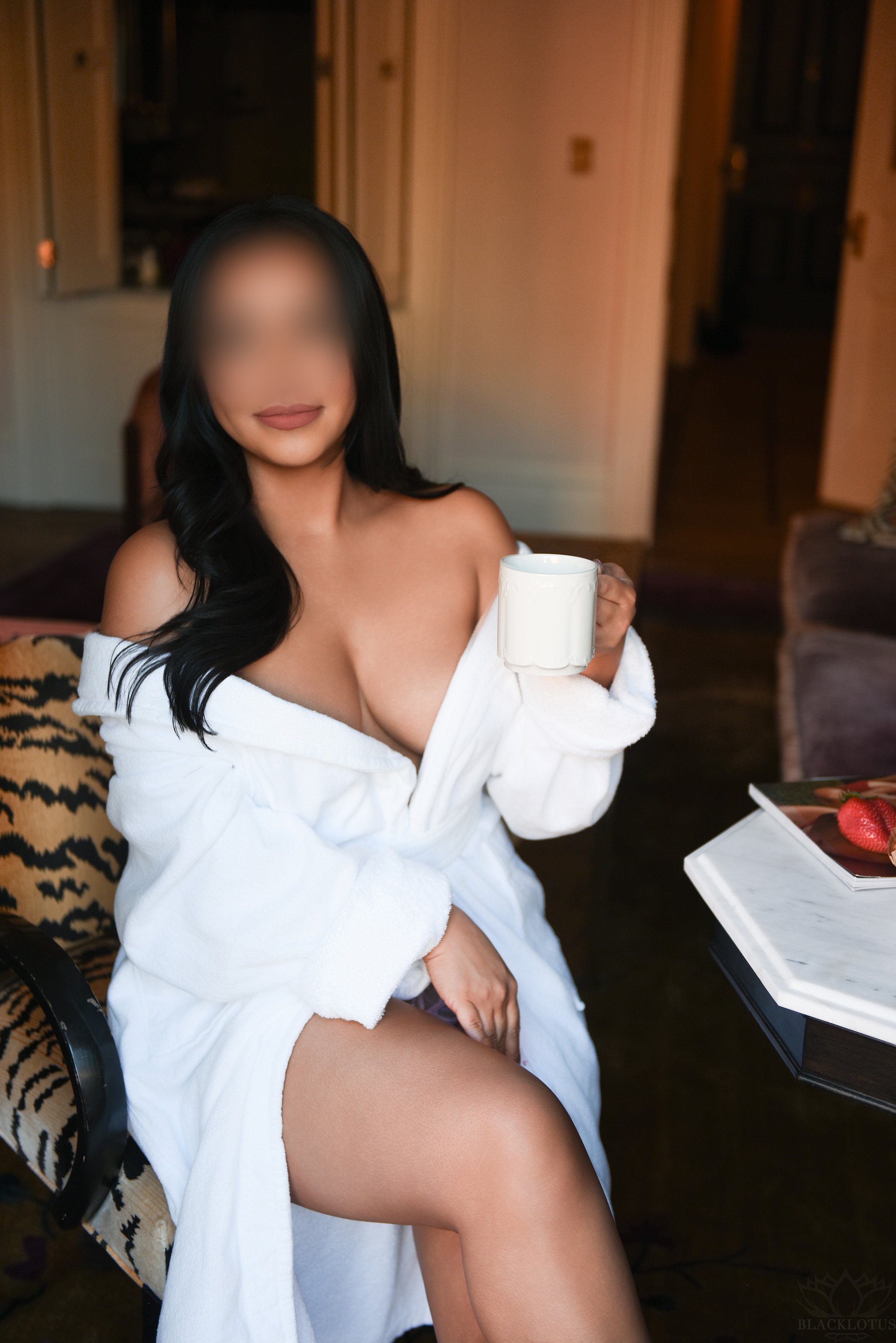 A woman with long dark hair, wrapped in a white towel, sitting on a chair in a cozy room, holding a white coffee mug, with a plate of strawberries on the table nearby.