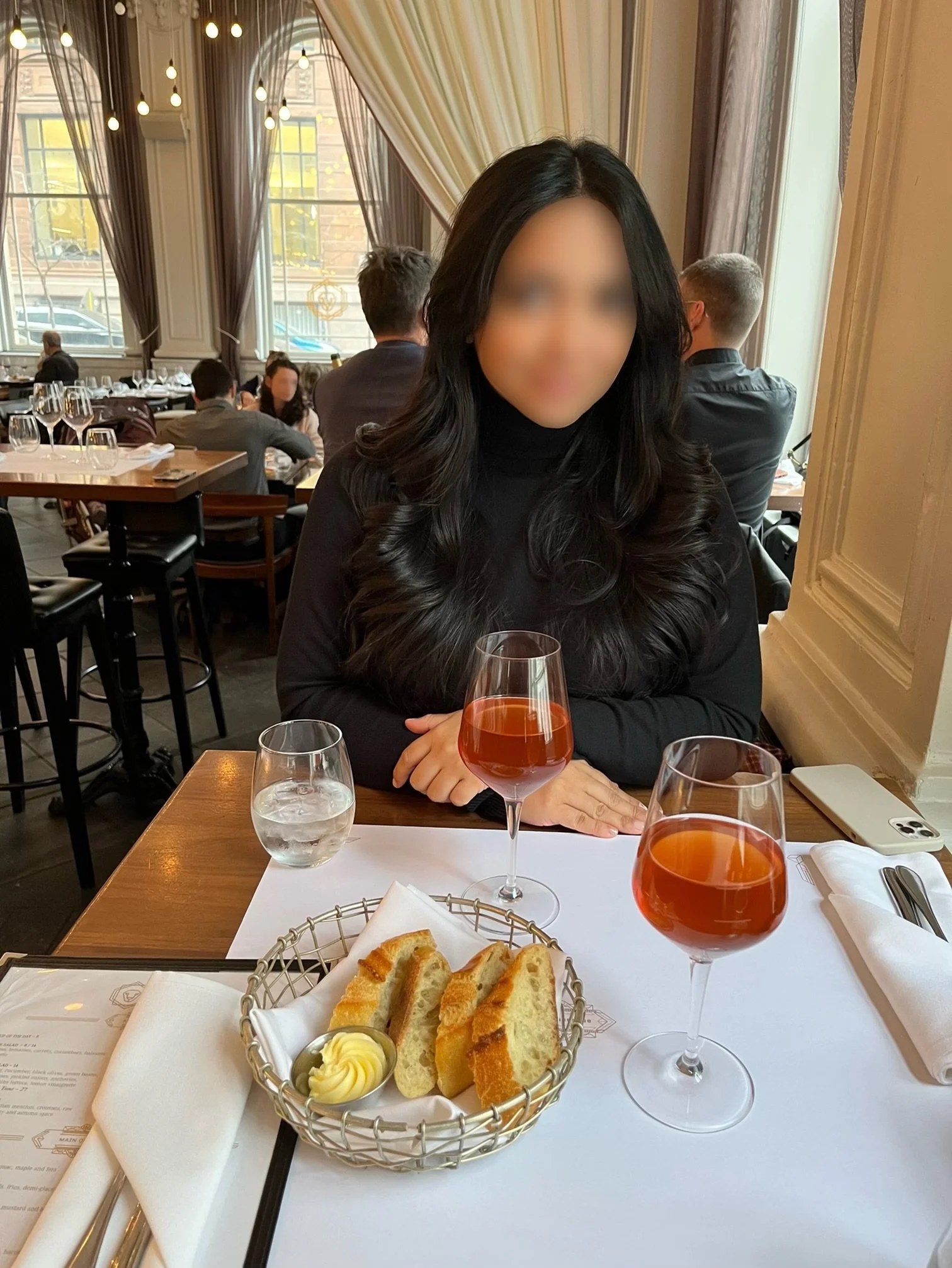A woman with long black hair and a black turtleneck sitting at a restaurant table with a glass of rosé wine. In the foreground, there is a basket with slices of bread, a small dish of butter, and a white napkin. The table is set with a water glass, s