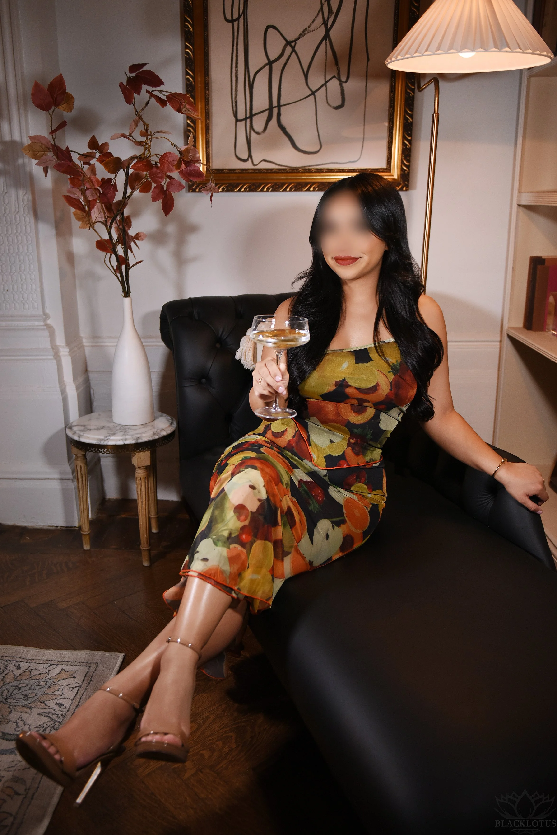 A woman with long dark hair and red lipstick sitting on a black leather sofa in a richly decorated room, holding a cocktail glass. She is wearing a colorful floral dress and high heels. Behind her is a tall white vase with red leaves, a marble side t
