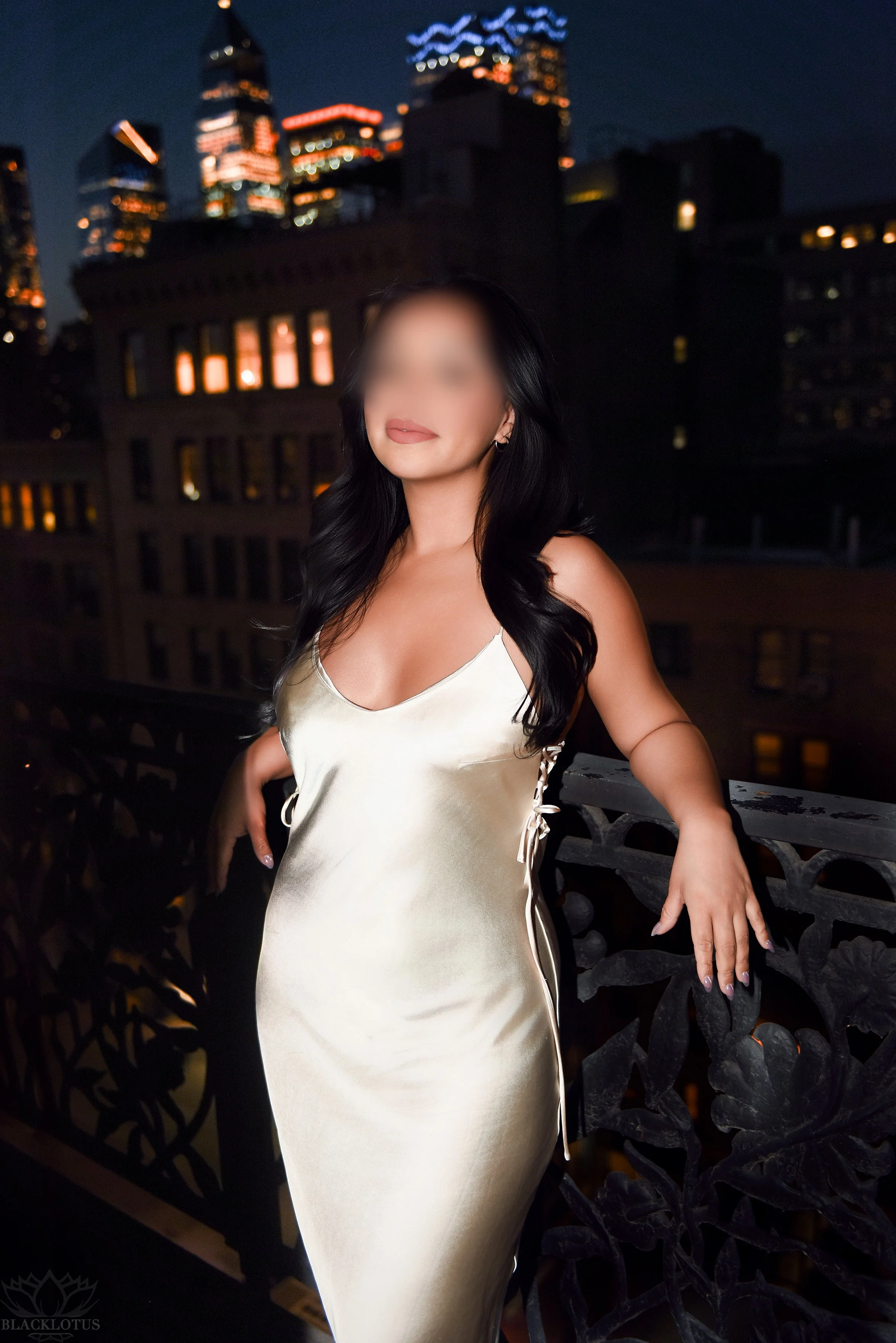 Amara with long hair wearing a shiny satin slip dress standing on a balcony at night with city buildings in the background.