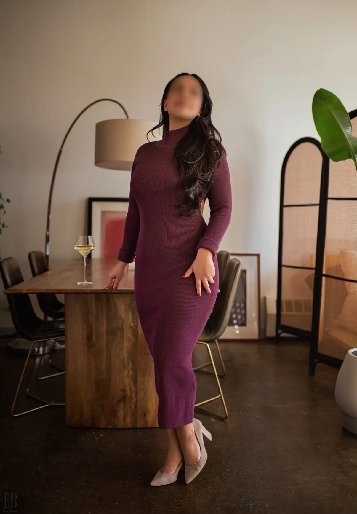 A woman in a form-fitting maroon turtleneck dress and beige high heels standing in a stylish dining room with a wine glass on a wooden table.