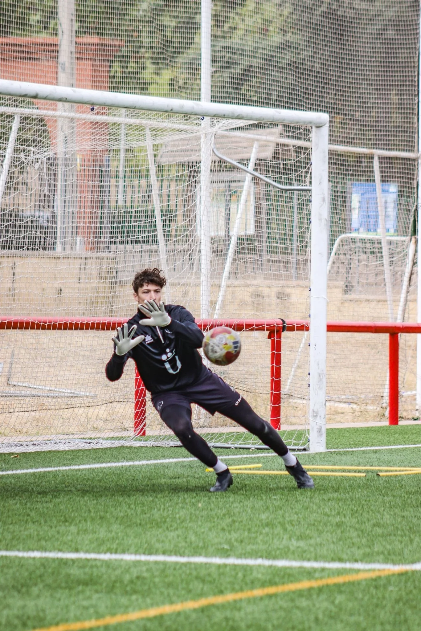 A soccer goalkeeper in black uniform, with gloves, is attempting to block a shot on goal during a game, inside a soccer field with green artificial turf, with a goalpost and net behind him.