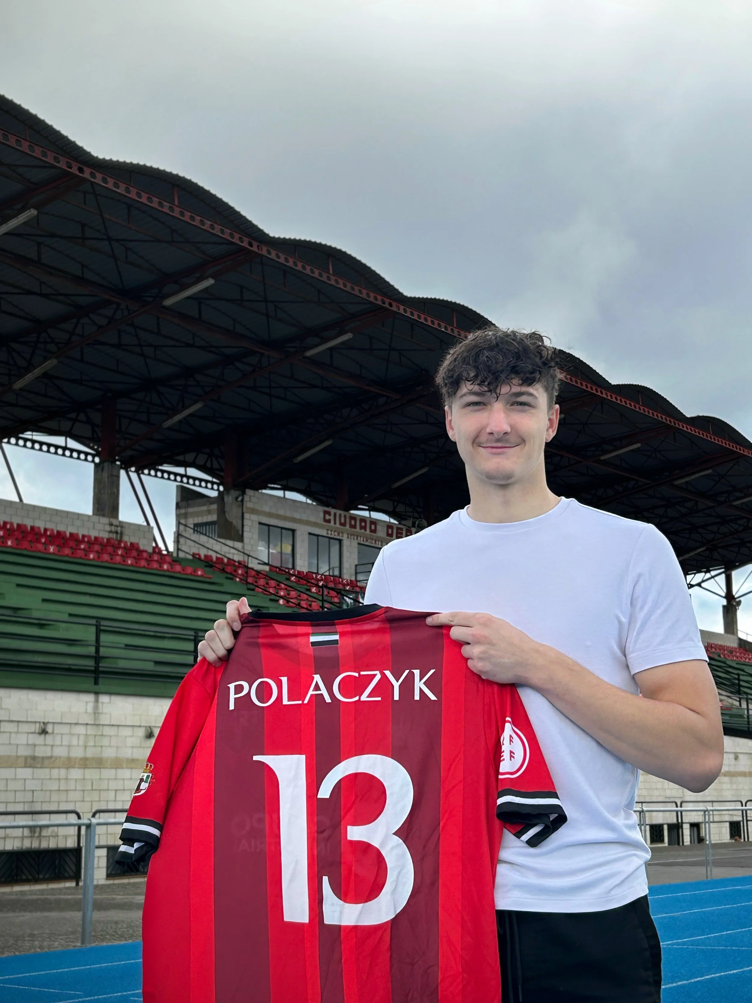 A young man with curly brown hair and a white t-shirt standing on a sports field, holding a red sports jersey with the name 'POLACZYK' and number 13, smiling at the camera with a stadium and cloudy sky in the background.