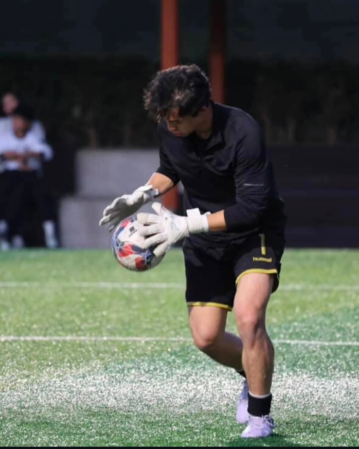 A soccer goalkeeper wearing a black uniform and white gloves catches a soccer ball on a field.