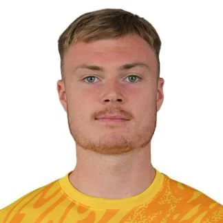 A young man with short light brown hair, light skin, and a slight beard, wearing a yellow sports jersey with orange patterns, standing against a plain white background.