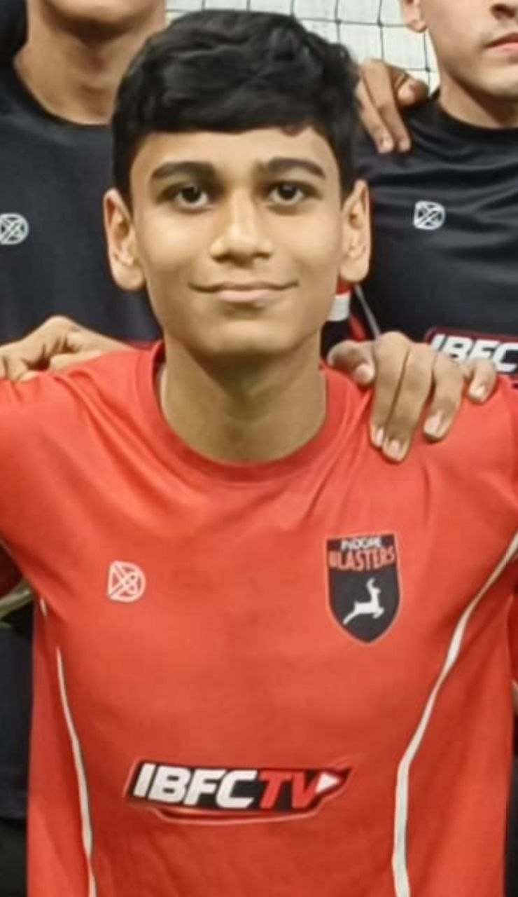 Young boy wearing a red sports jersey with a logo that says "Power Laste" and "IBFCTV". He has short dark hair and is smiling at the camera. Two other individuals are partially visible behind him, with one resting a hand on his shoulder.
