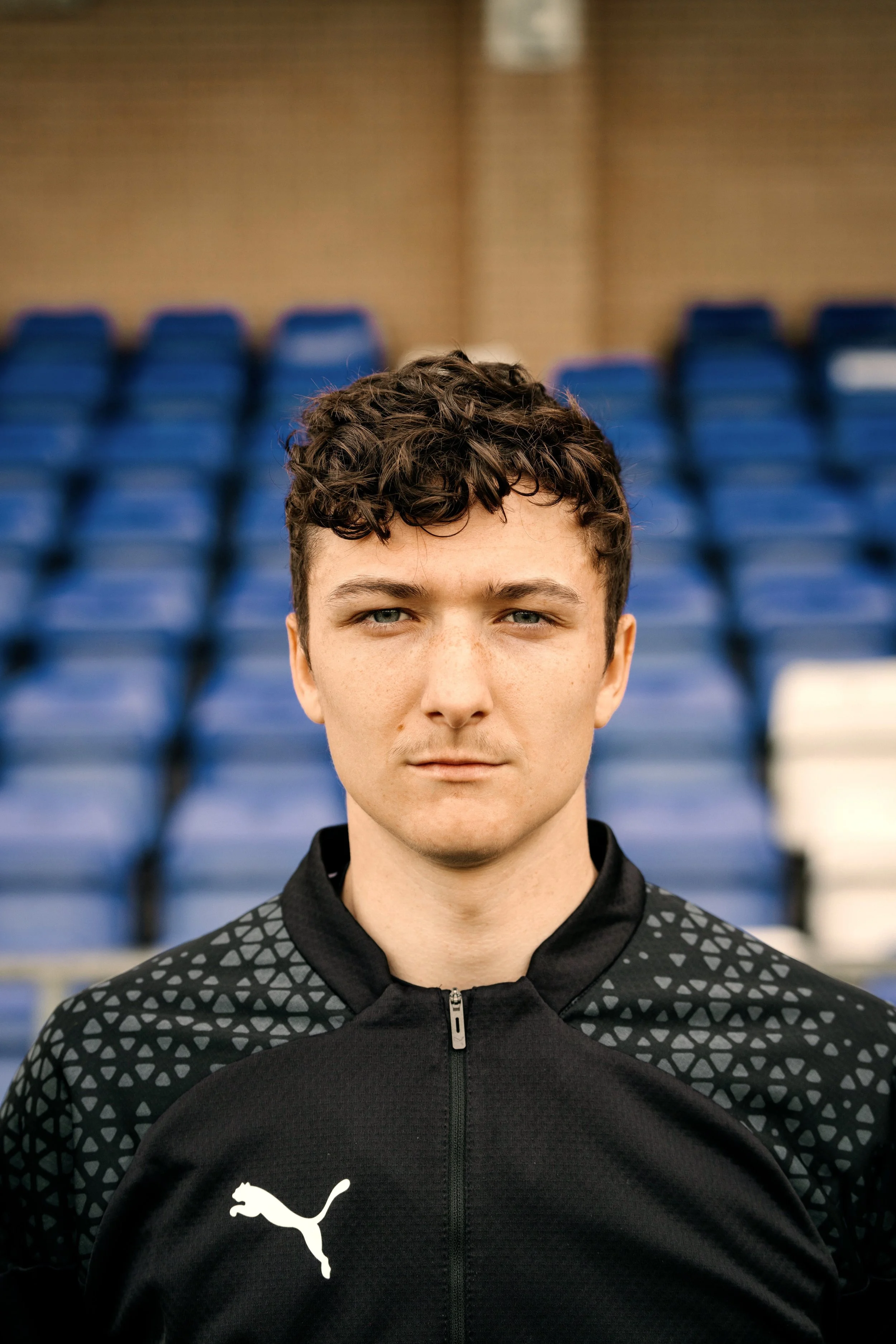 A young man with curly hair wearing a black Puma sports jacket with geometric pattern on the shoulders, standing in front of empty blue stadium seats.