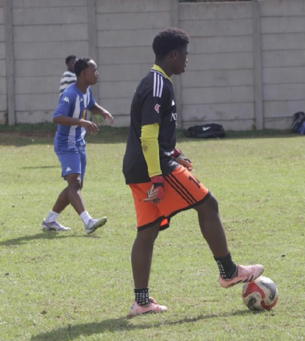 A young soccer player in a black and yellow uniform with orange shorts is controlling a soccer ball on a grass field. There are other players and a concrete fence in the background.