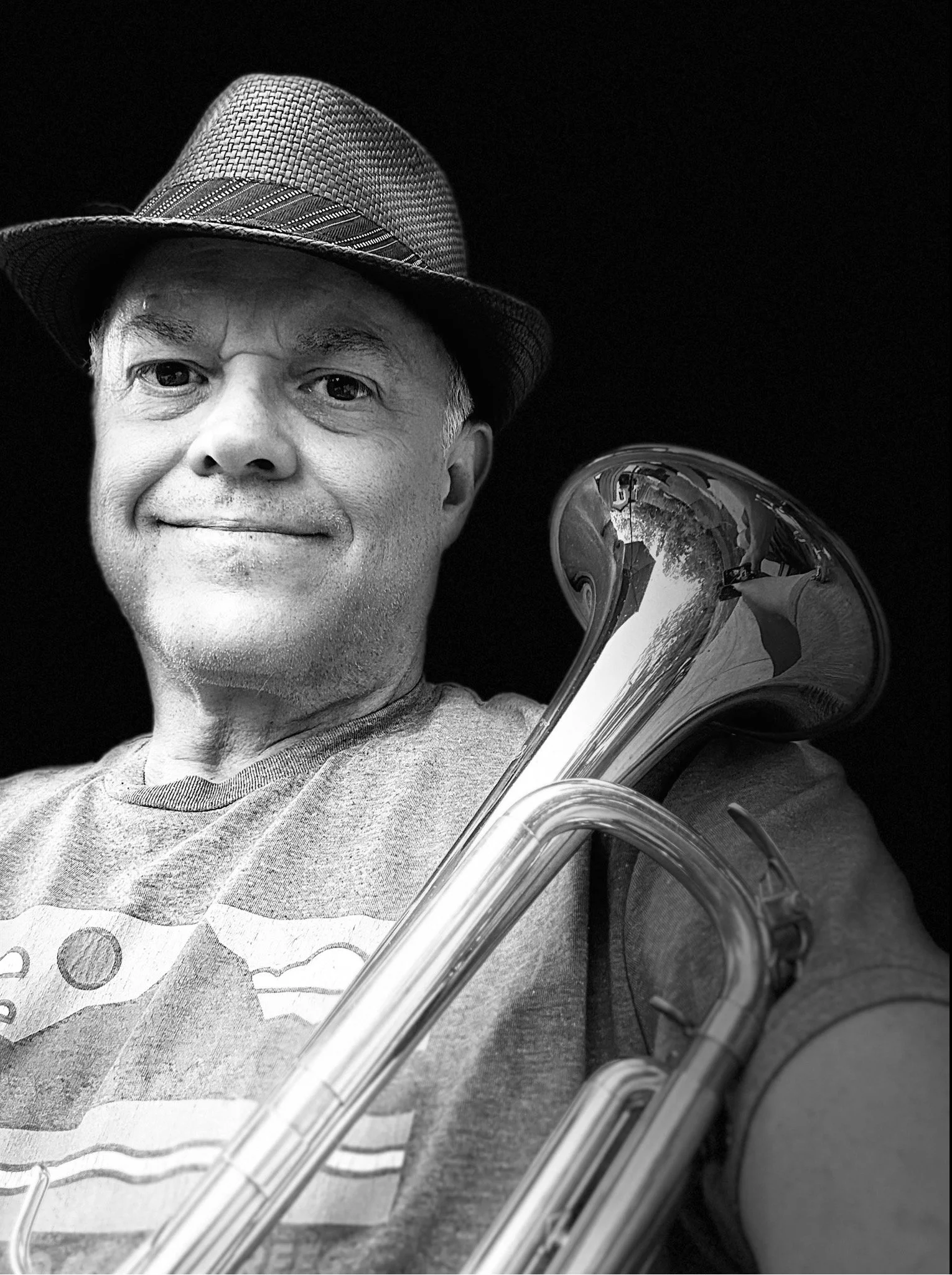 A man smiling, wearing a fedora hat and polka dot shirt, holding a shiny brass musical instrument, possibly a trumpet, with reflections visible on the instrument's surface.