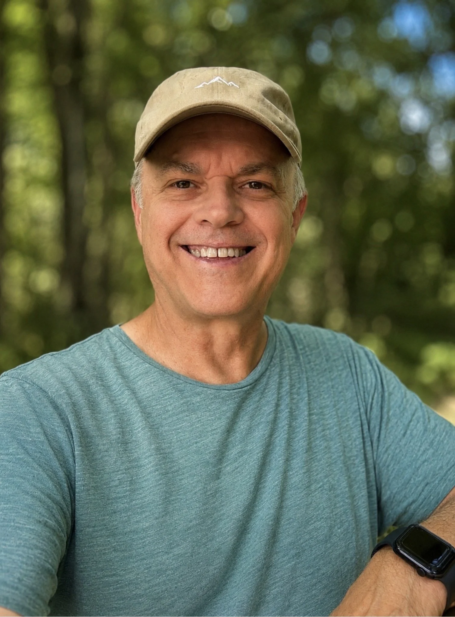 Close-up of a smiling man wearing a tan cap and a teal t-shirt, with trees in the blurred background.