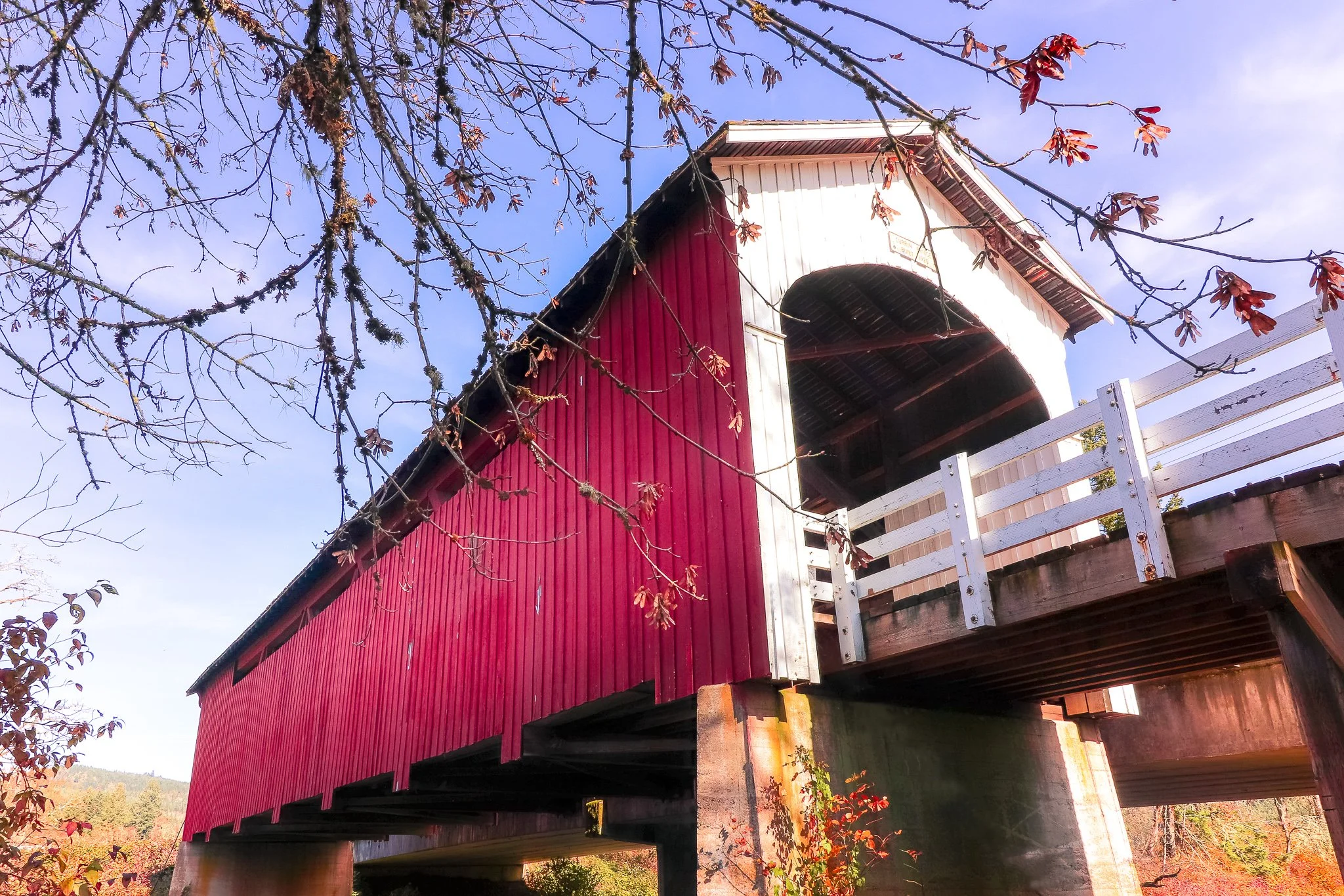 A red covered bridge with white accents, elevated on wooden supports, and surrounded by trees with autumn foliage and a clear blue sky.