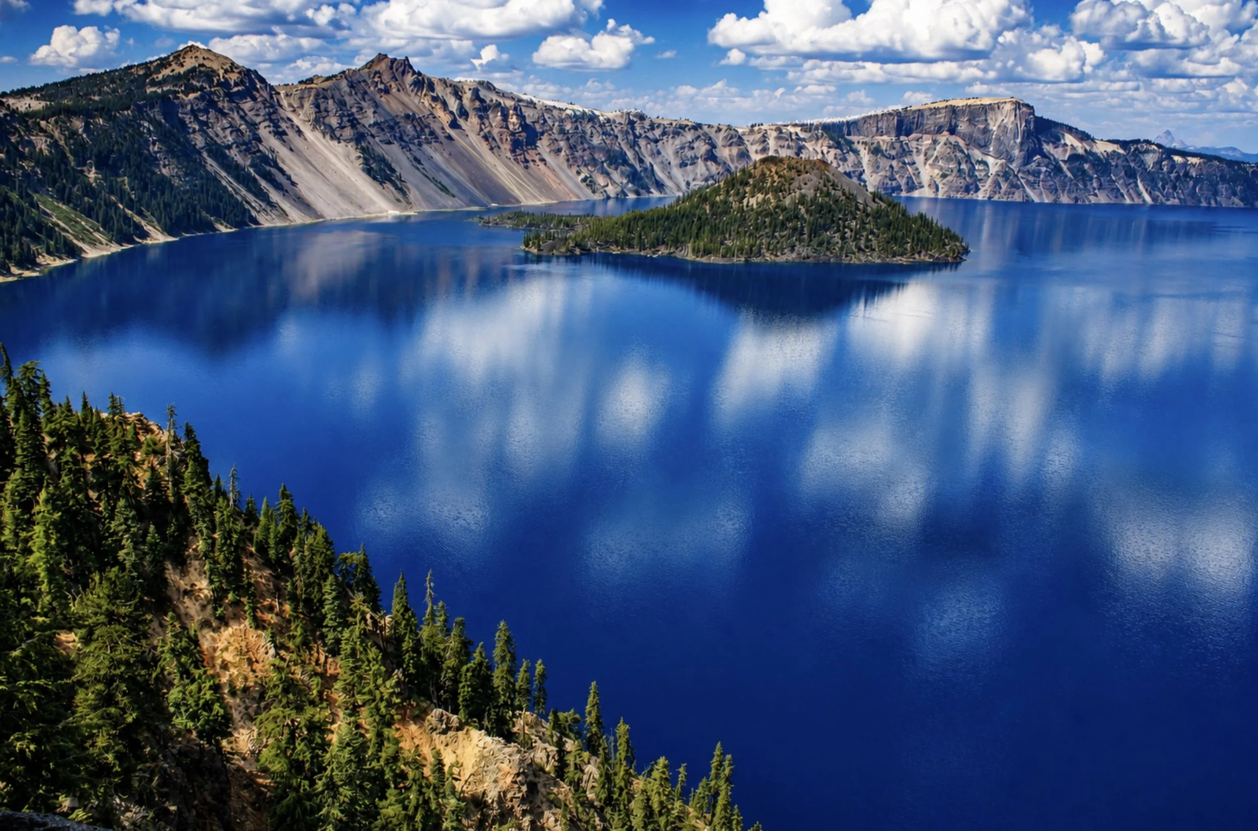 A large blue lake surrounded by forested hills and rugged mountains under a partly cloudy sky.