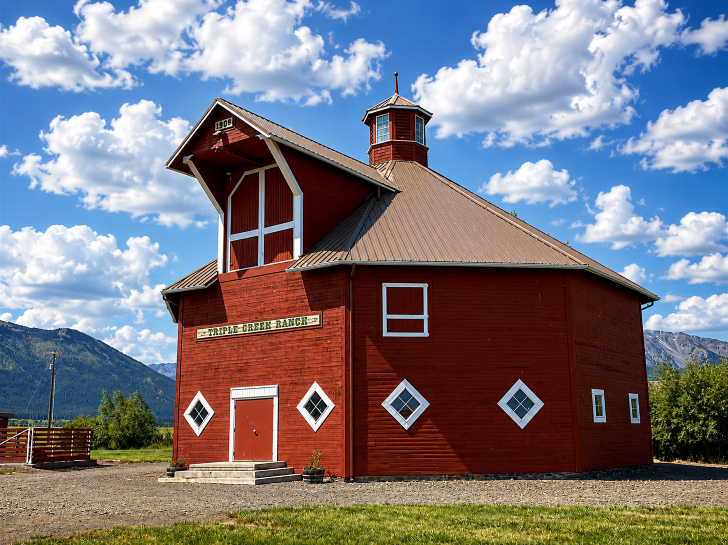 Red barn with gooseneck windows, diamond-shaped window accents, and a sign that reads 'TRIPLE CREEK RANCH,' set against a backdrop of mountains and a partly cloudy sky.