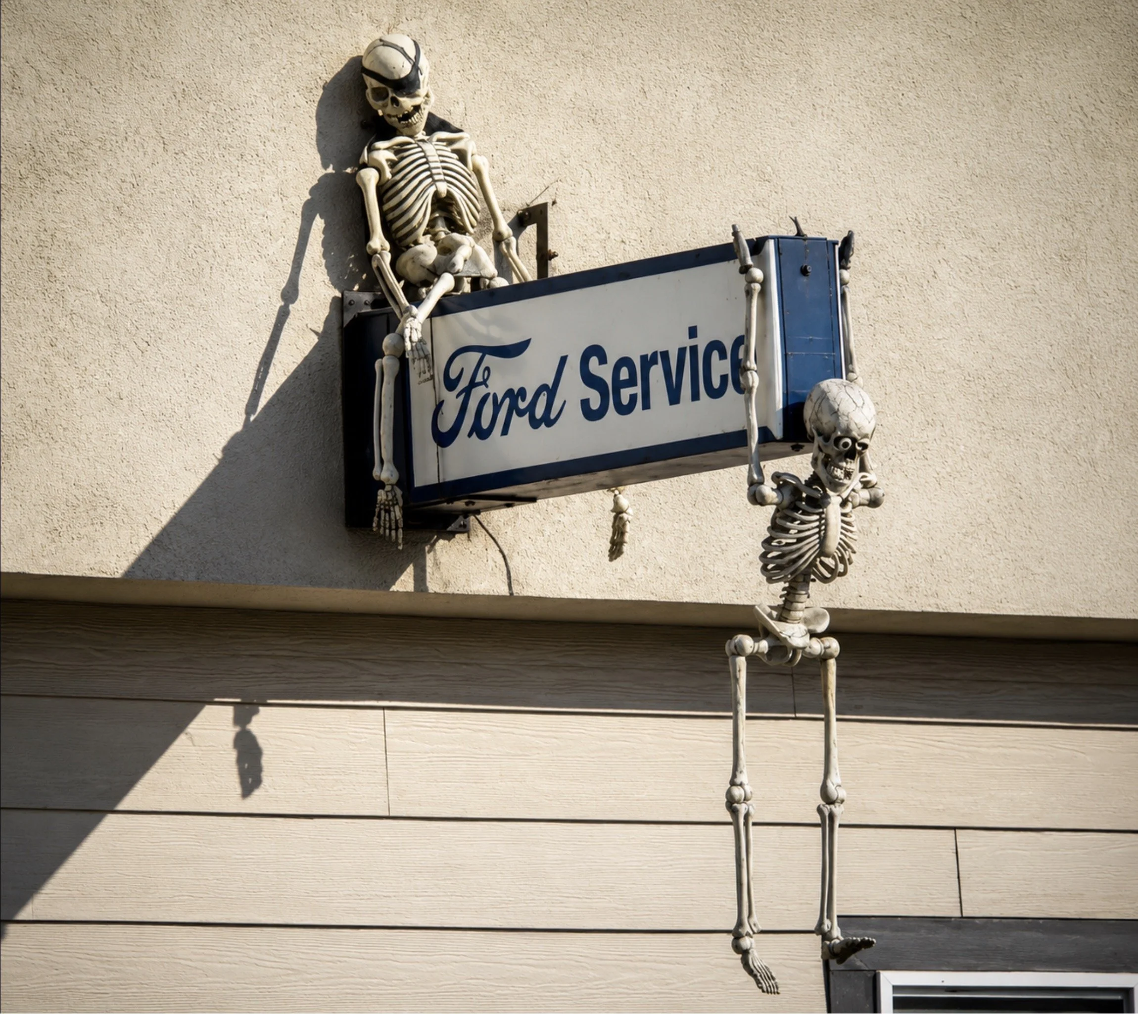 Skeleton seated on a shelf under a skeleton hanging vertically, both attached to a wall with a 'Ford Service' sign.