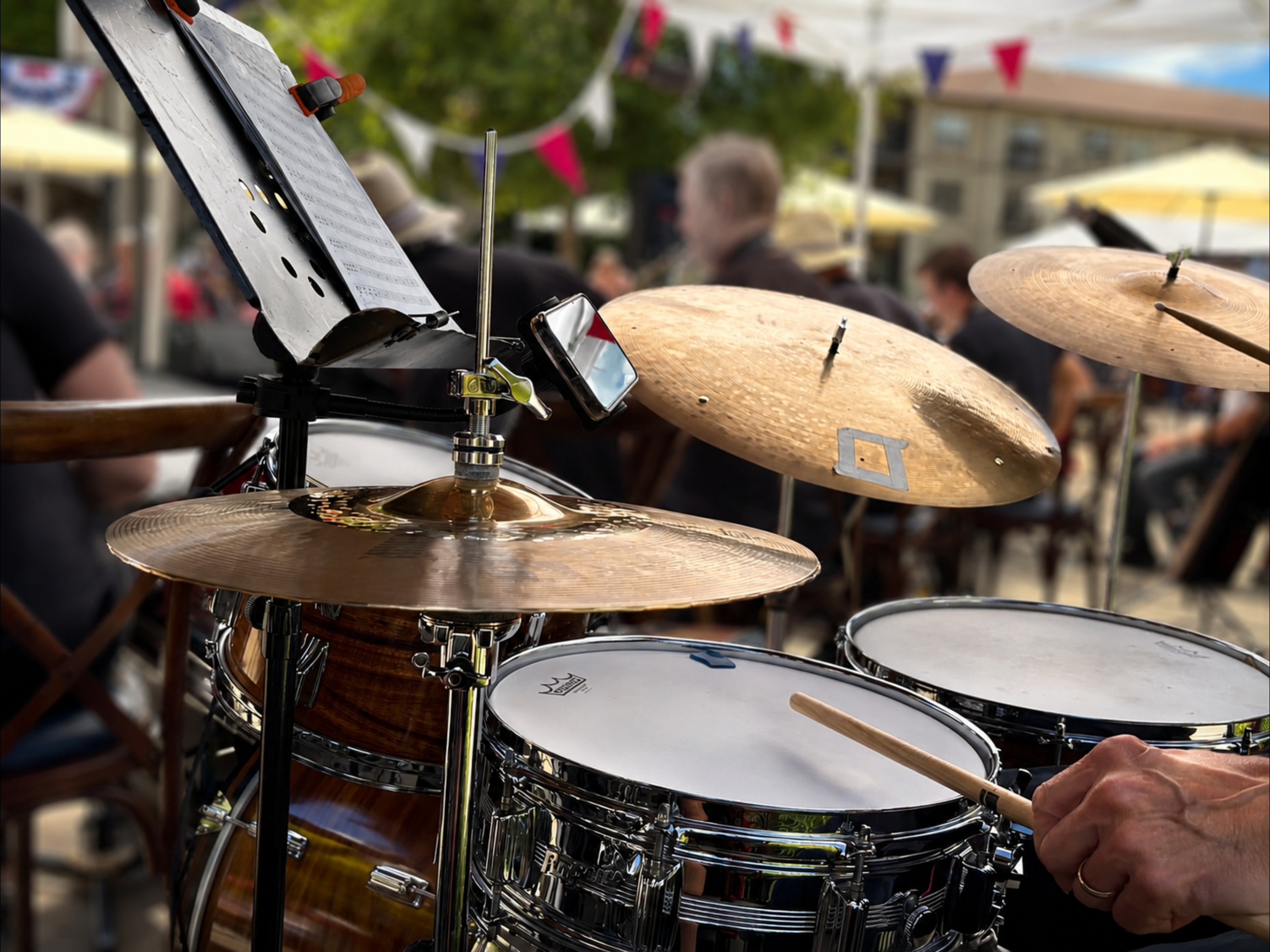 Close-up of a drum set with cymbals and a music stand at an outdoor event, with people blurred in the background.