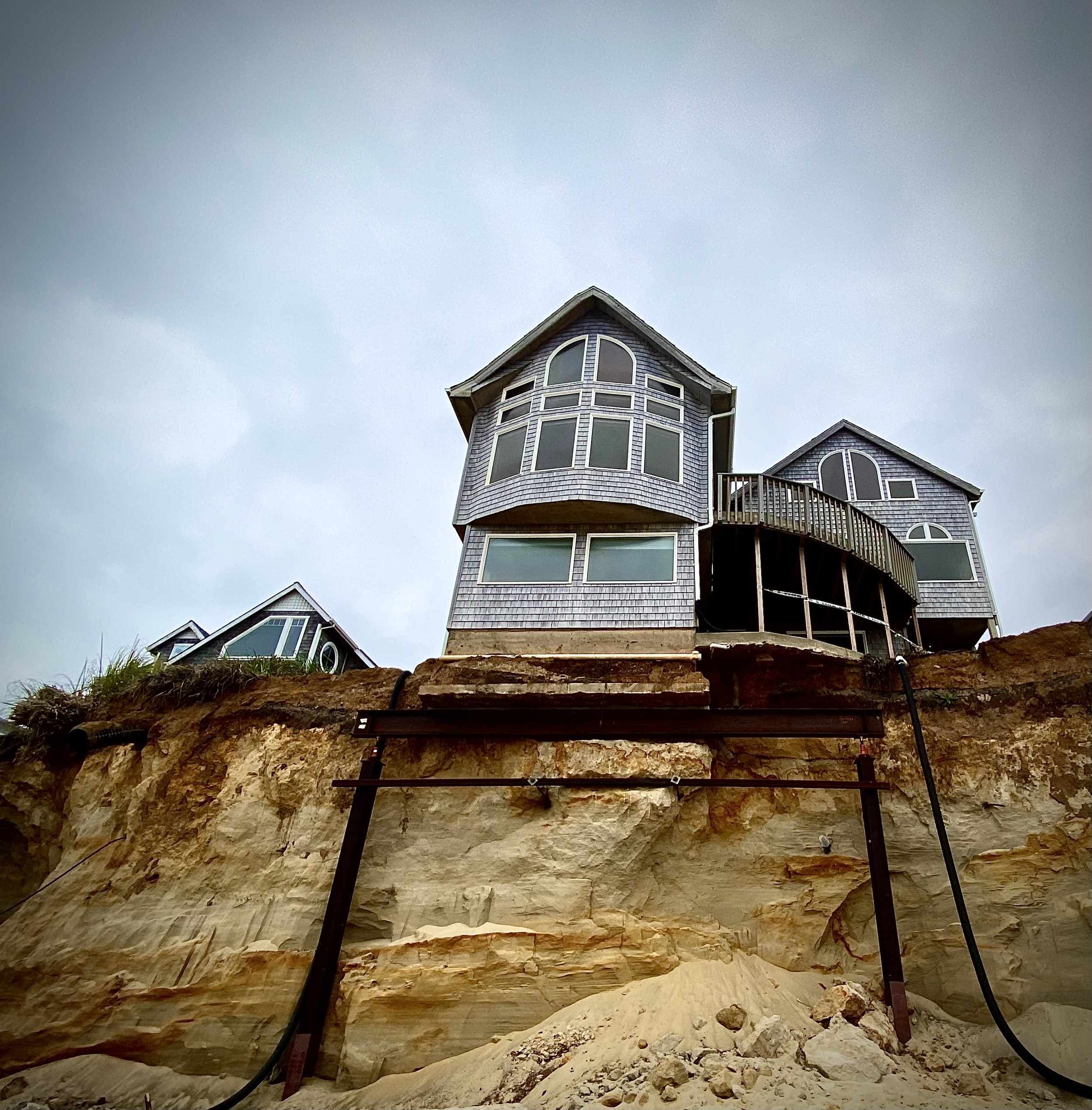Under construction house with a steep wooden roof, large windows, and a balcony, built on a rocky hill with construction materials and scaffolding in the foreground.