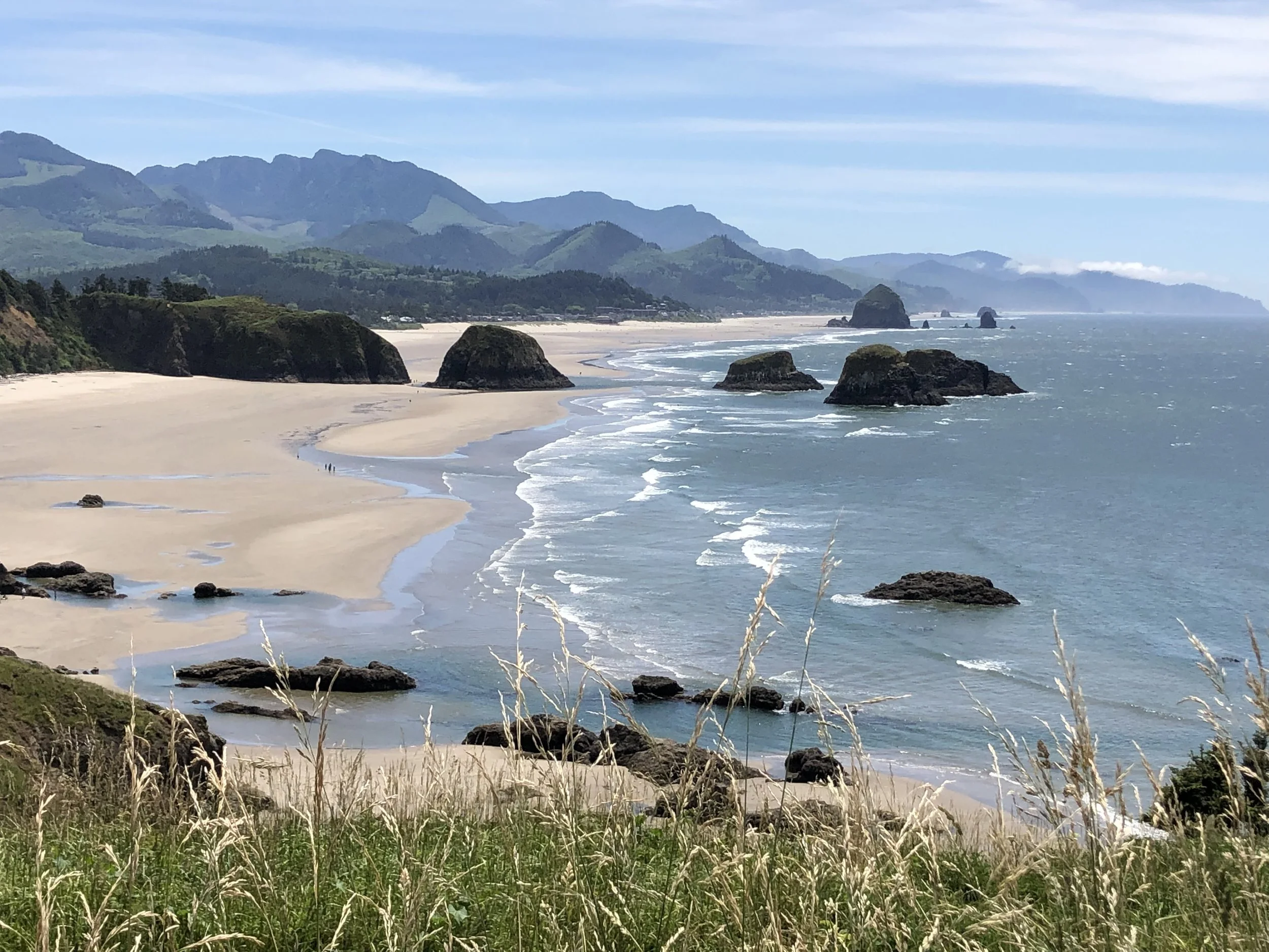 Coastal landscape with sandy beach, large rocks in the water, and green mountains in the background under a partly cloudy sky.