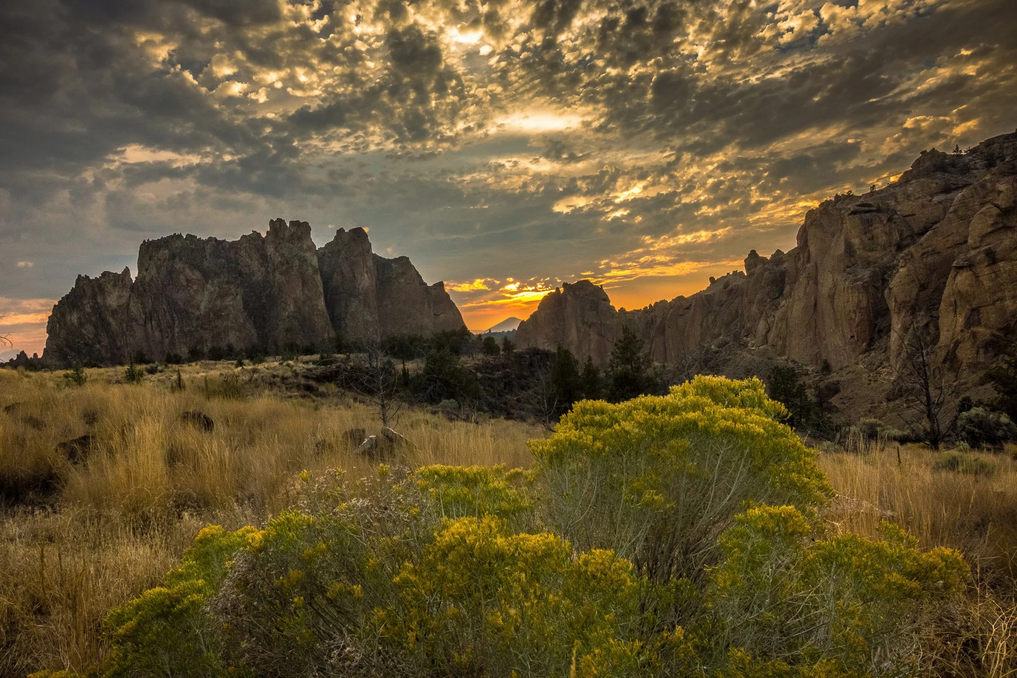 A scenic landscape at sunset featuring rugged rocky mountains, a sky filled with scattered clouds illuminated by the setting sun, and desert vegetation in the foreground.