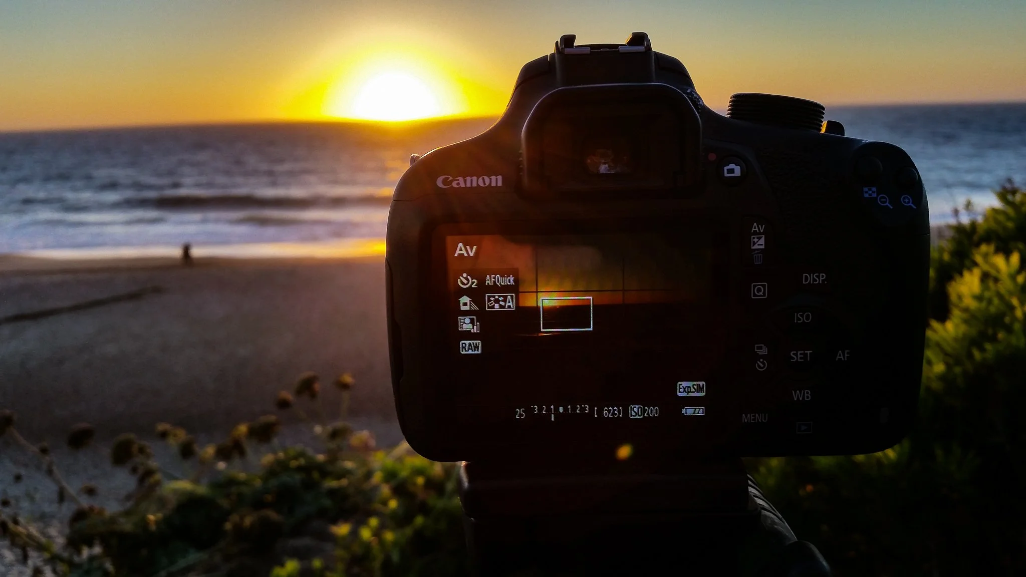 Close-up of a Canon camera's display screen capturing a sunset over the ocean at the beach with a person walking in the distance.