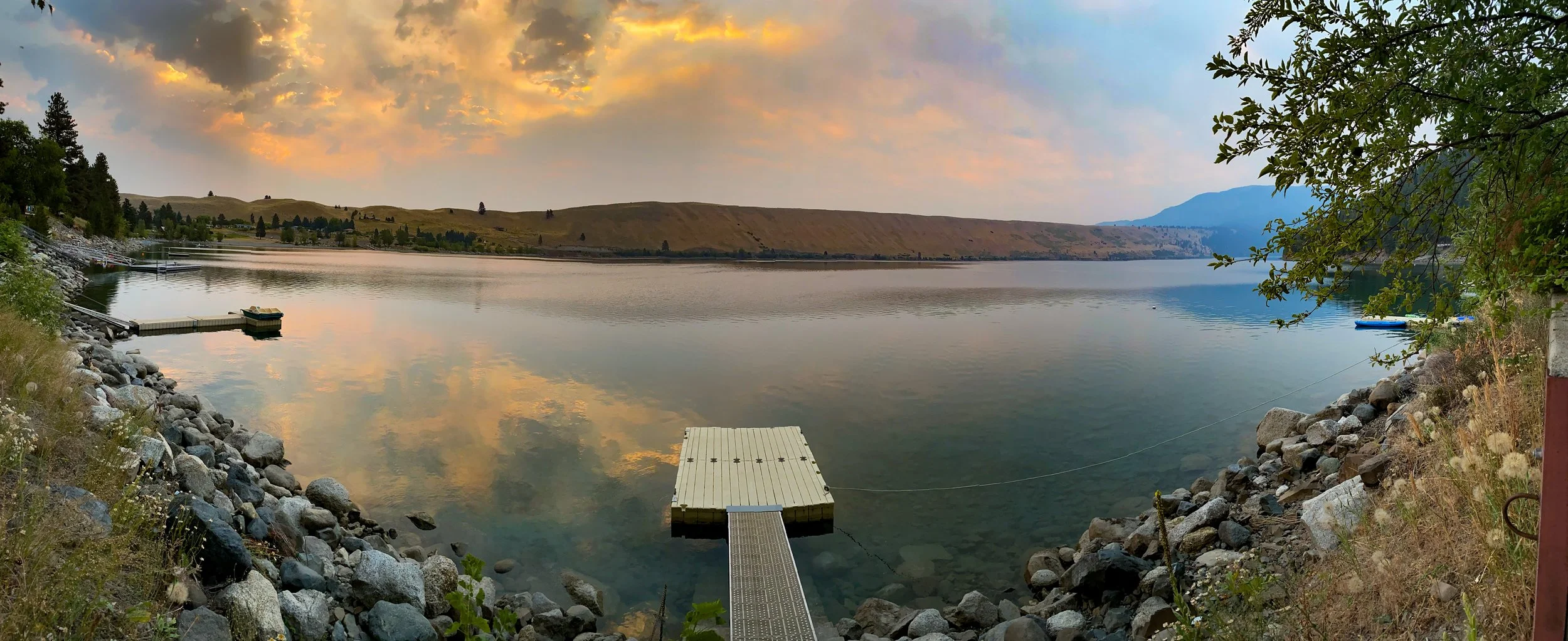 A peaceful lake scene during sunset with calm water reflecting clouds and sky, surrounded by rocky shoreline, trees, and distant hills.