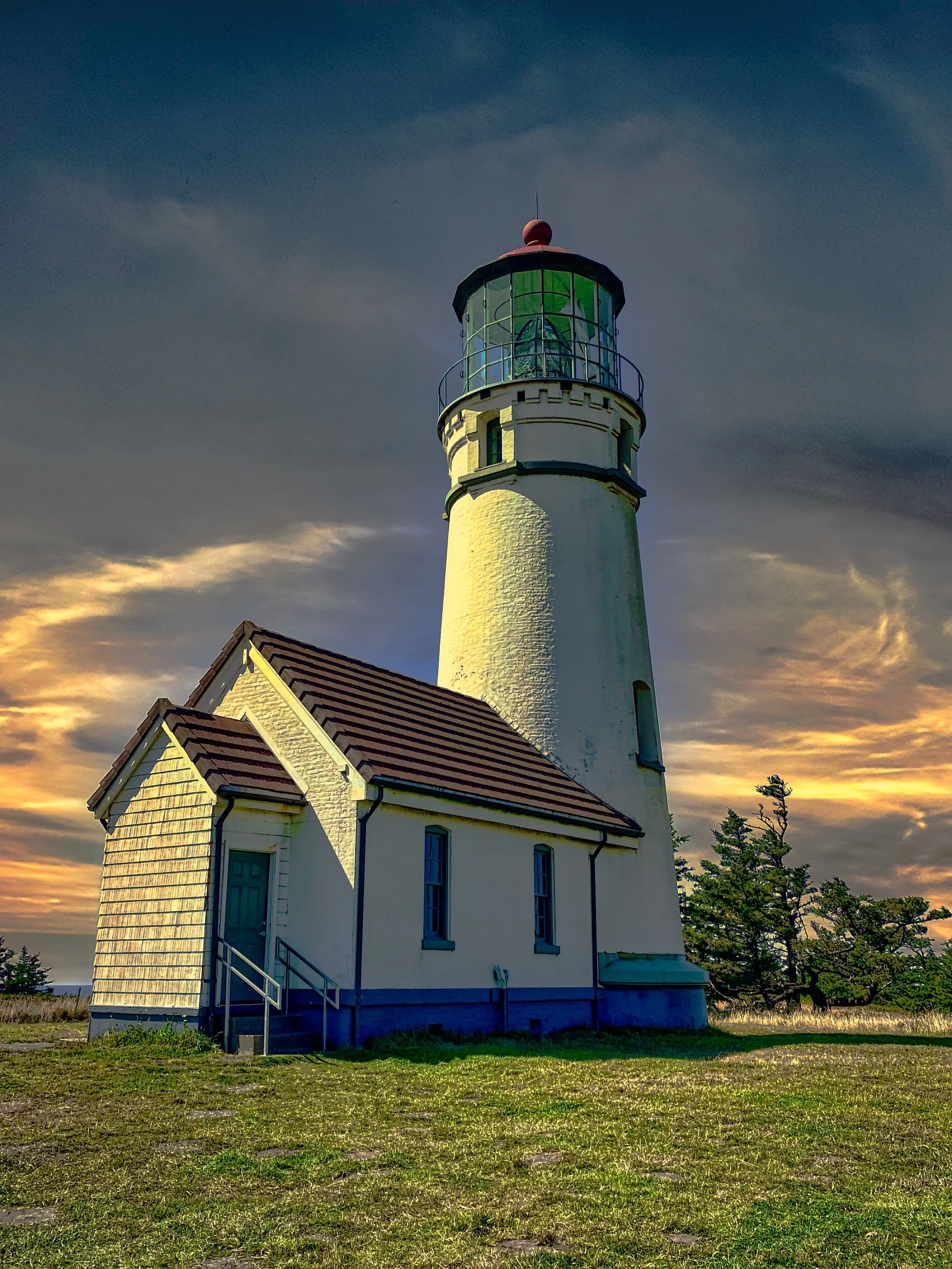 A white lighthouse with a red top, attached to a small building with a stairs leading up to a side door, set against a sky with clouds during sunset or sunrise.