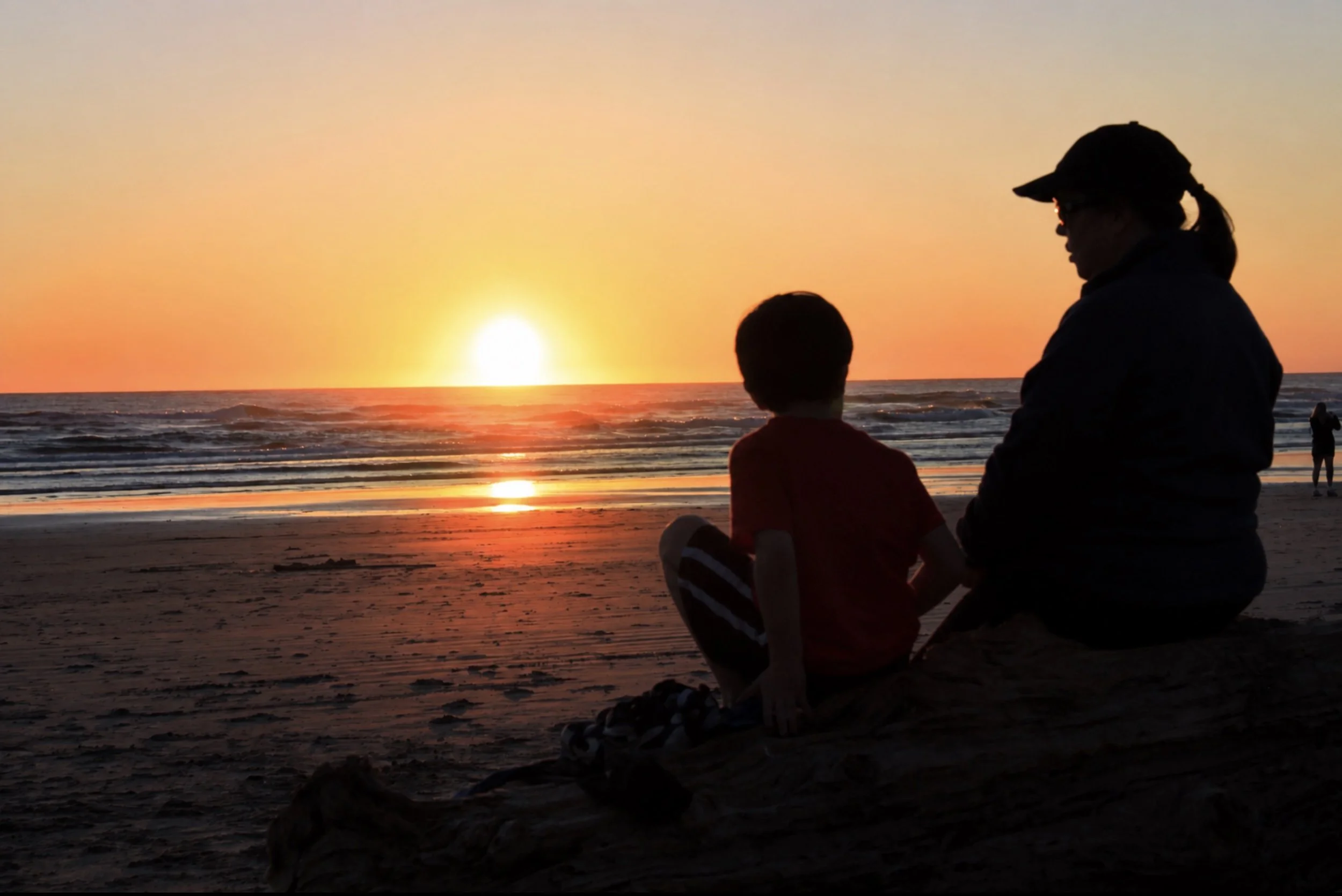 A silhouetted woman and child sitting on the beach at sunset, with the ocean in the background. The woman is wearing a cap and glasses, and there are other people walking along the shoreline.