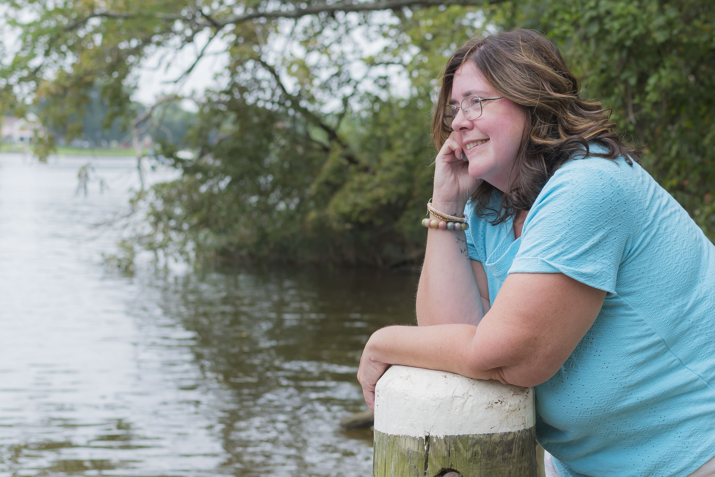 A woman with glasses and long, wavy brown hair sitting by a body of water, smiling, wearing a light blue blouse, with trees in the background.