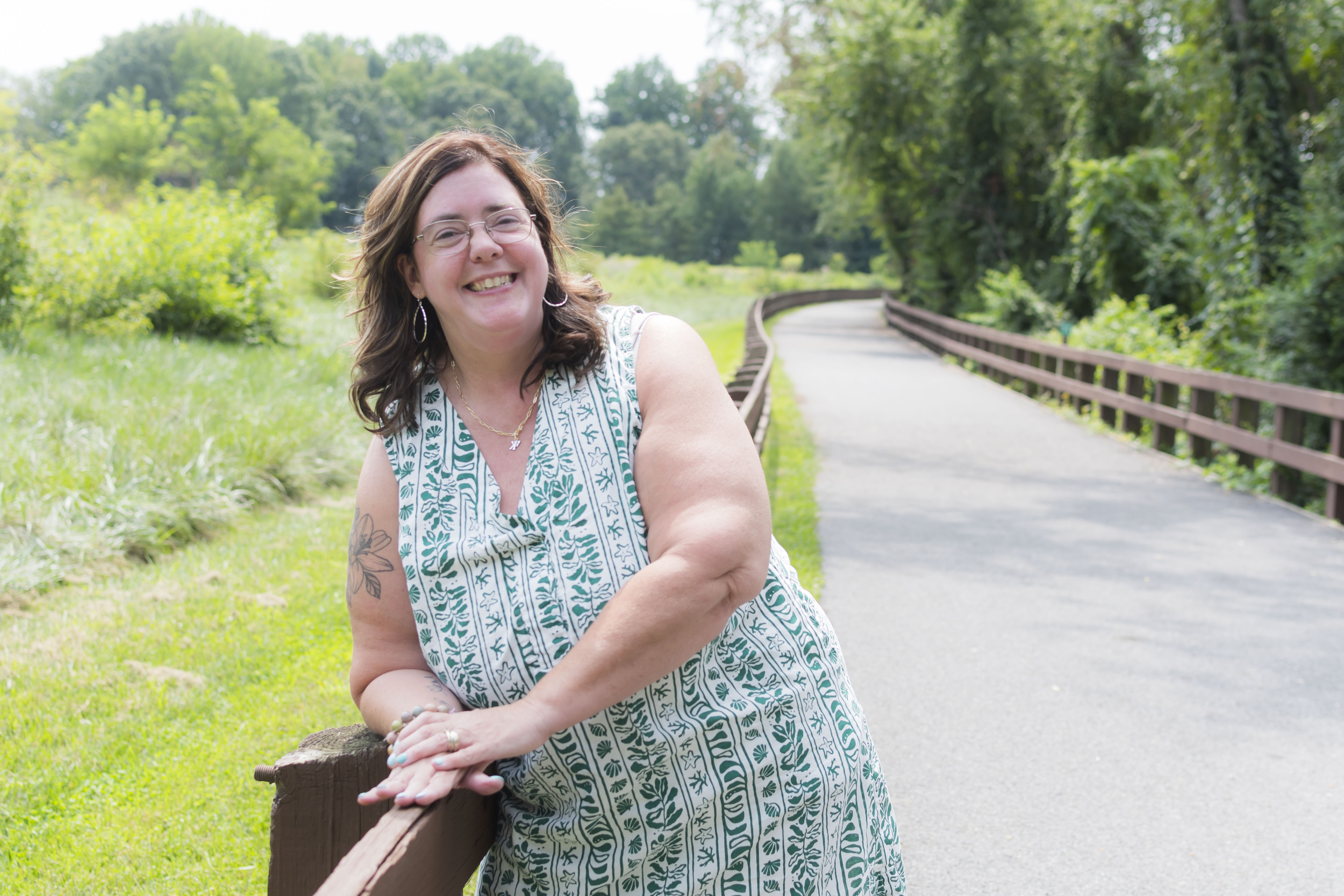 A smiling woman with glasses and long brown hair, wearing a patterned sleeveless dress, leaning on a wooden fence in a lush green park with a winding path and dense trees in the background.