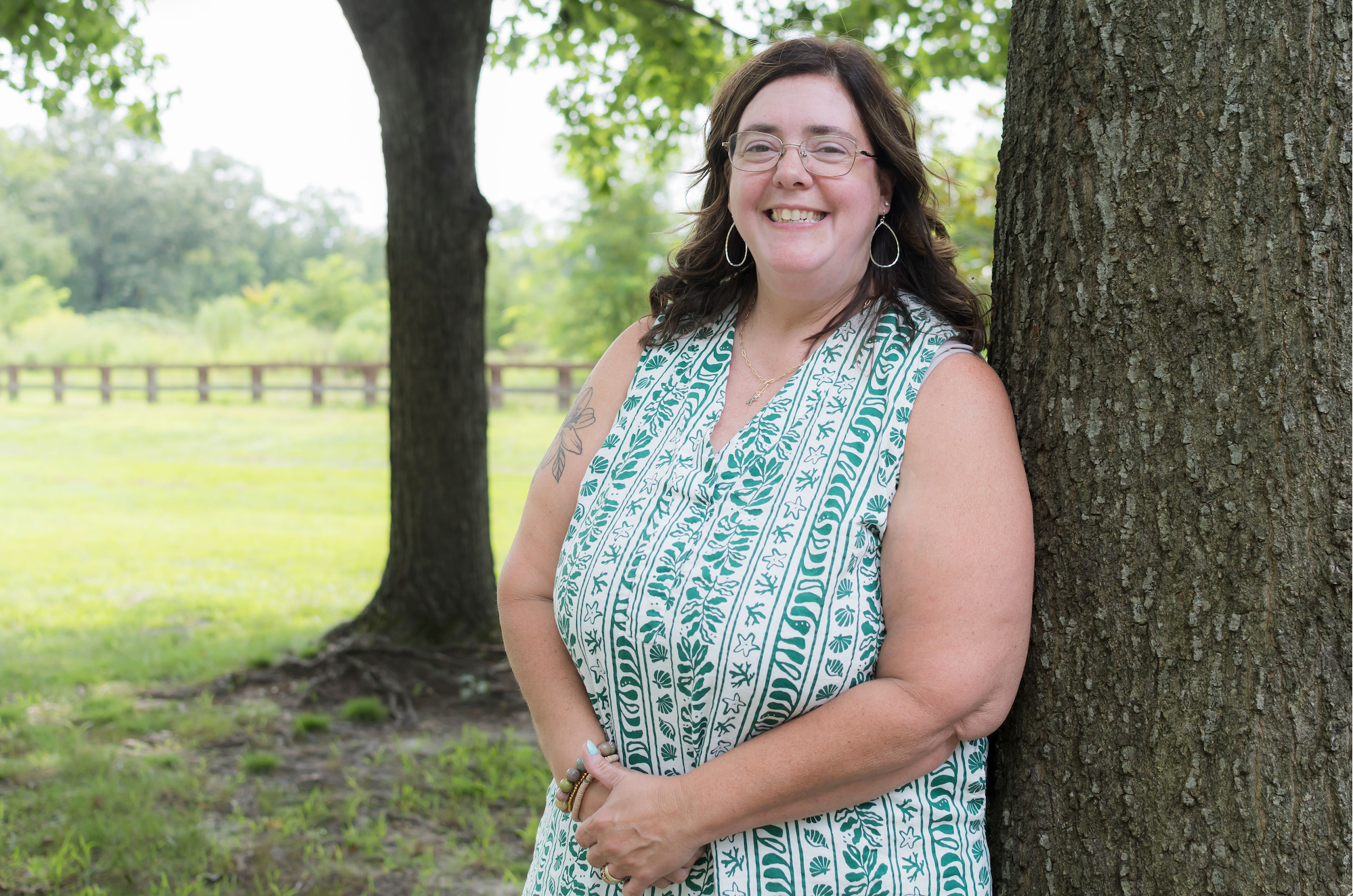 A smiling woman with glasses and earrings standing outdoors next to a tree, wearing a sleeveless patterned dress with green and white designs, with green trees and a fence in the background.