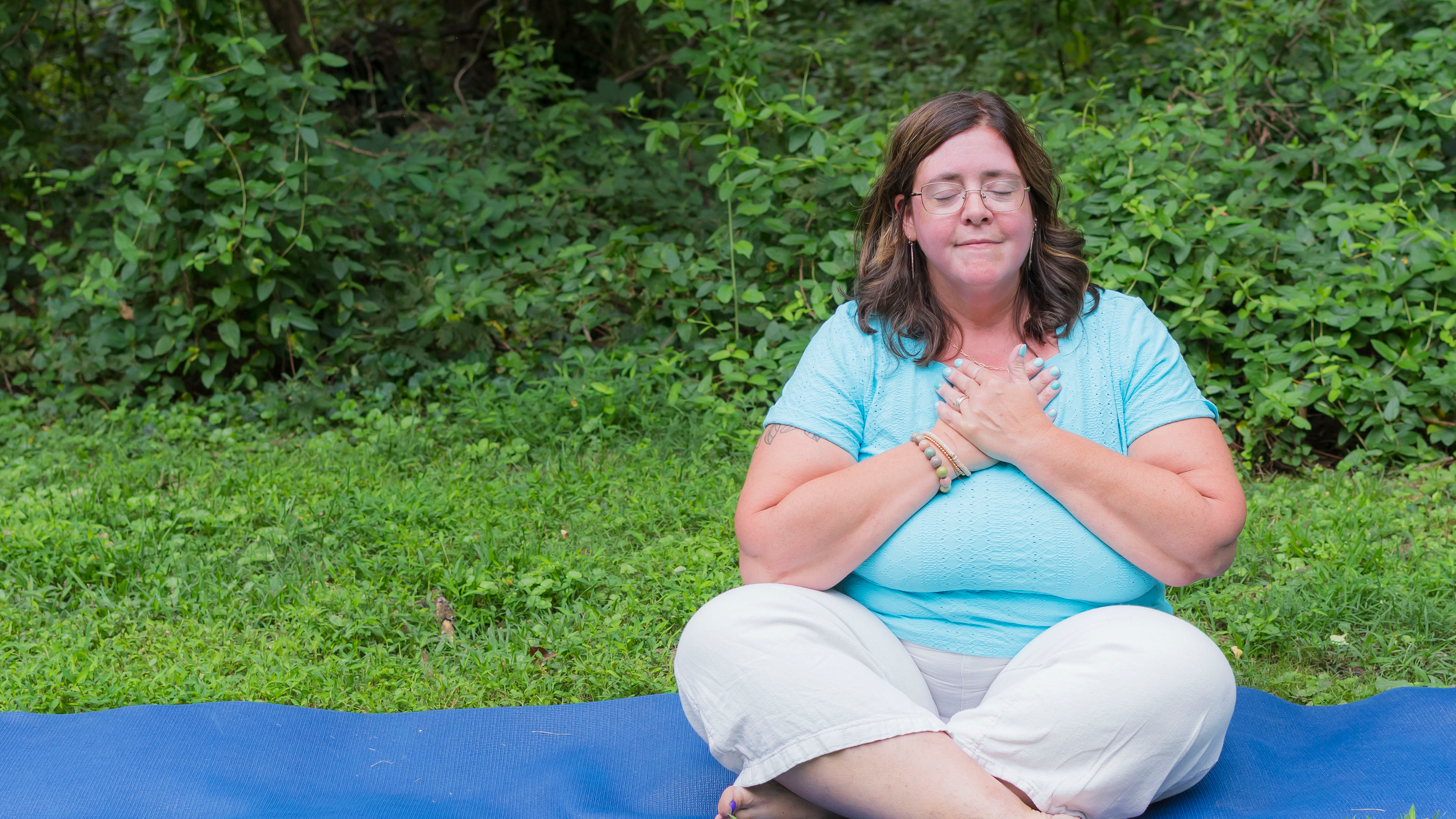 A woman with shoulder-length brown hair, glasses, and wearing a light blue shirt, is sitting outdoors on a blue yoga mat with her eyes closed and hands over her chest, in a meditative pose. She is surrounded by green foliage and grass.